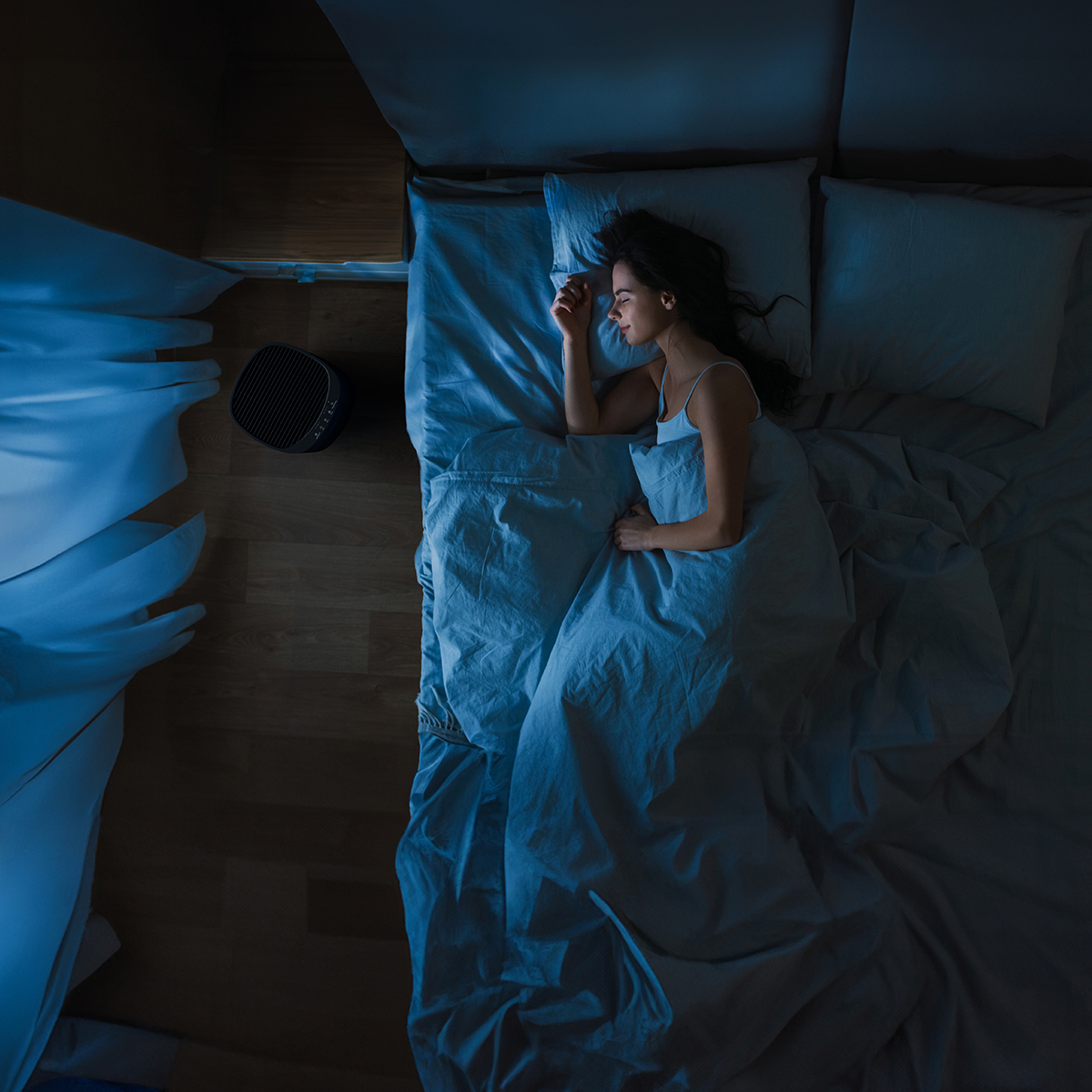 Une femme dort sous une couette bleue dans le noir. Un purificateur d'air au sol.