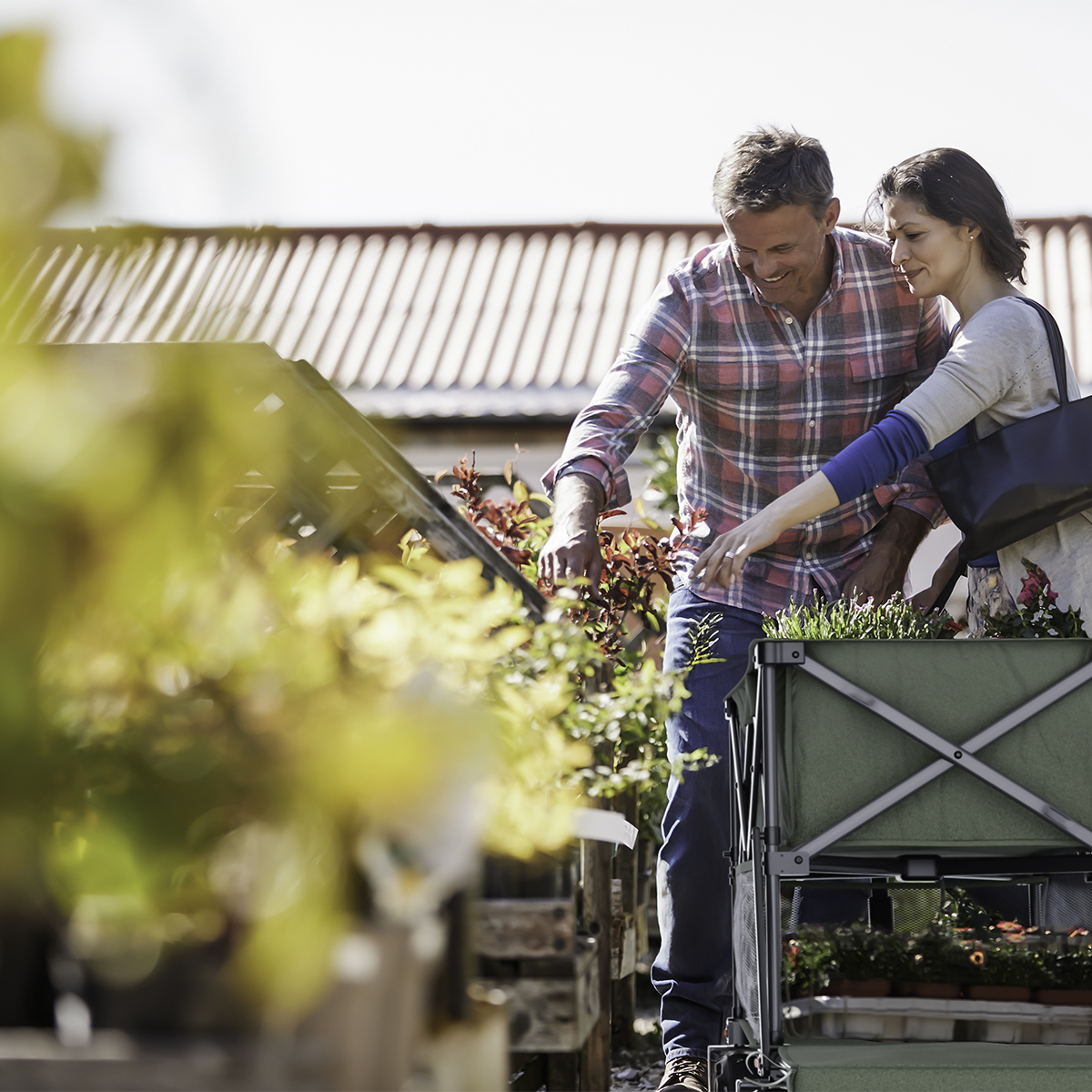 Couple shops for plants at a garden center, with a green cart full of flowers.