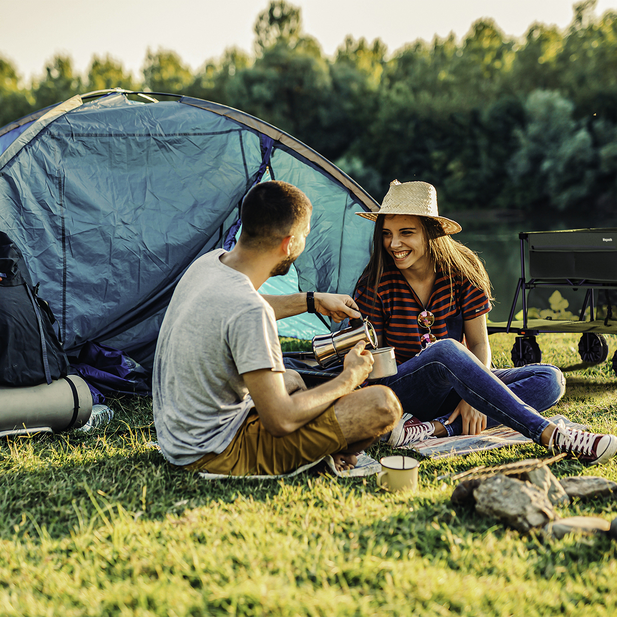 Couple camping by a lake. Man pours coffee for smiling woman. Tent, cart, trees.