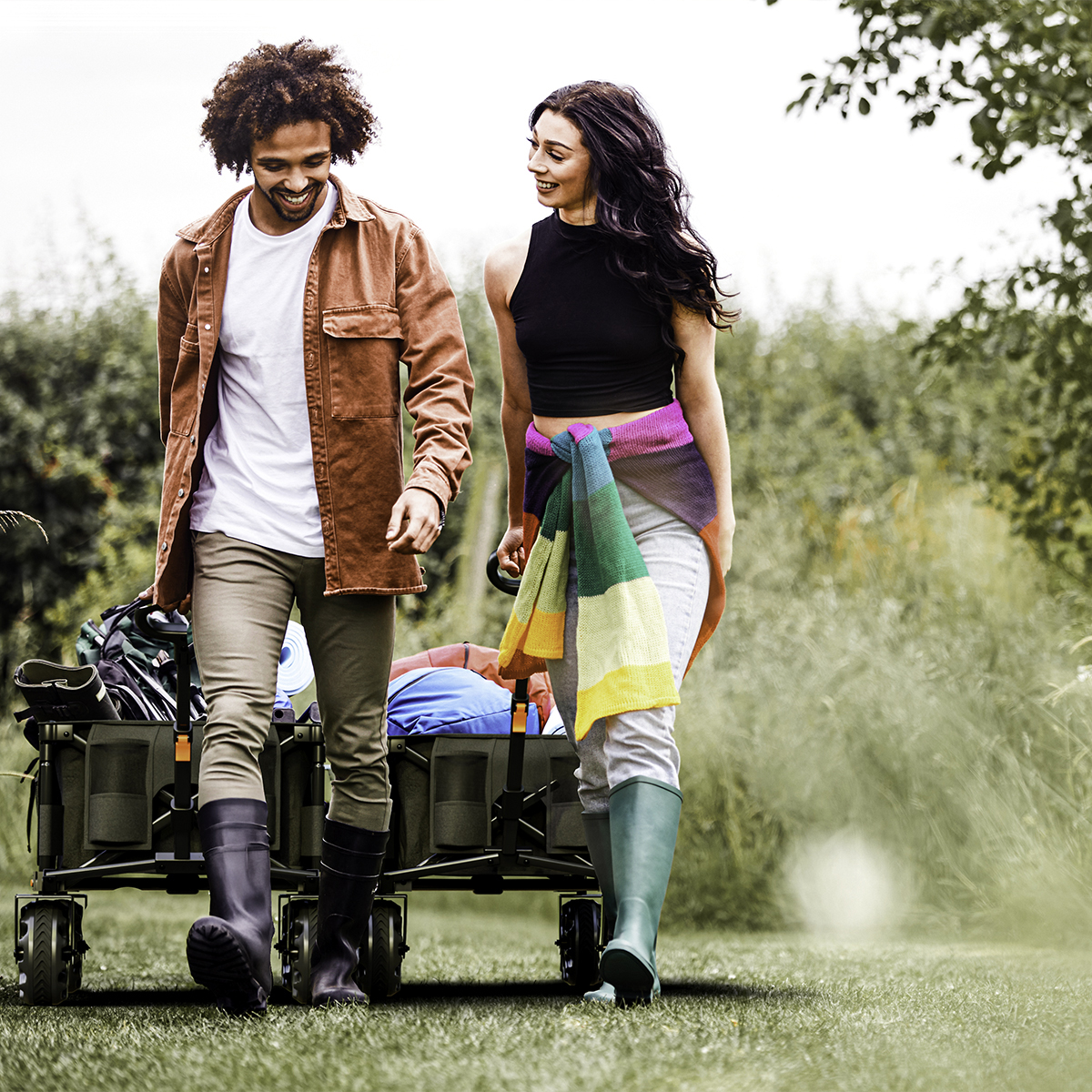 Man and woman pull a wagon with gear through green grass. Cloudy sky.