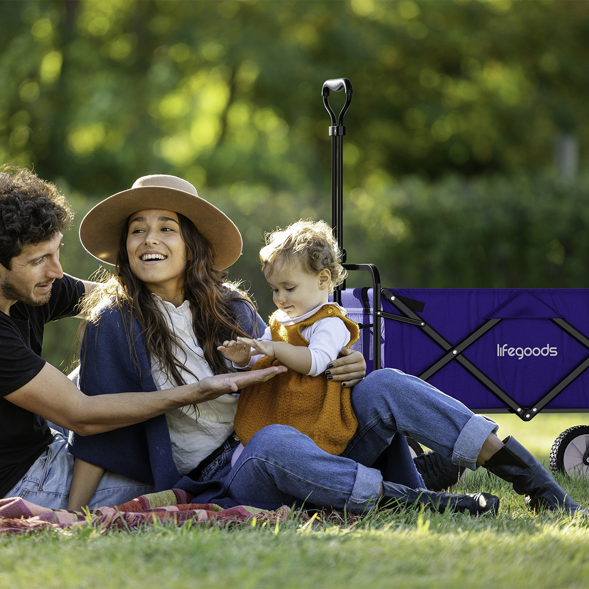 A man, woman, and baby sit on a blanket in grass, next to a blue wagon.
