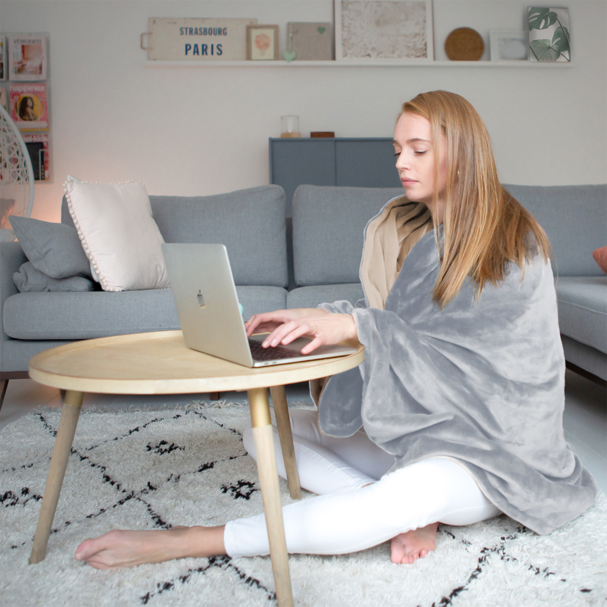 A woman wearing a blanket sits on the floor using a laptop.