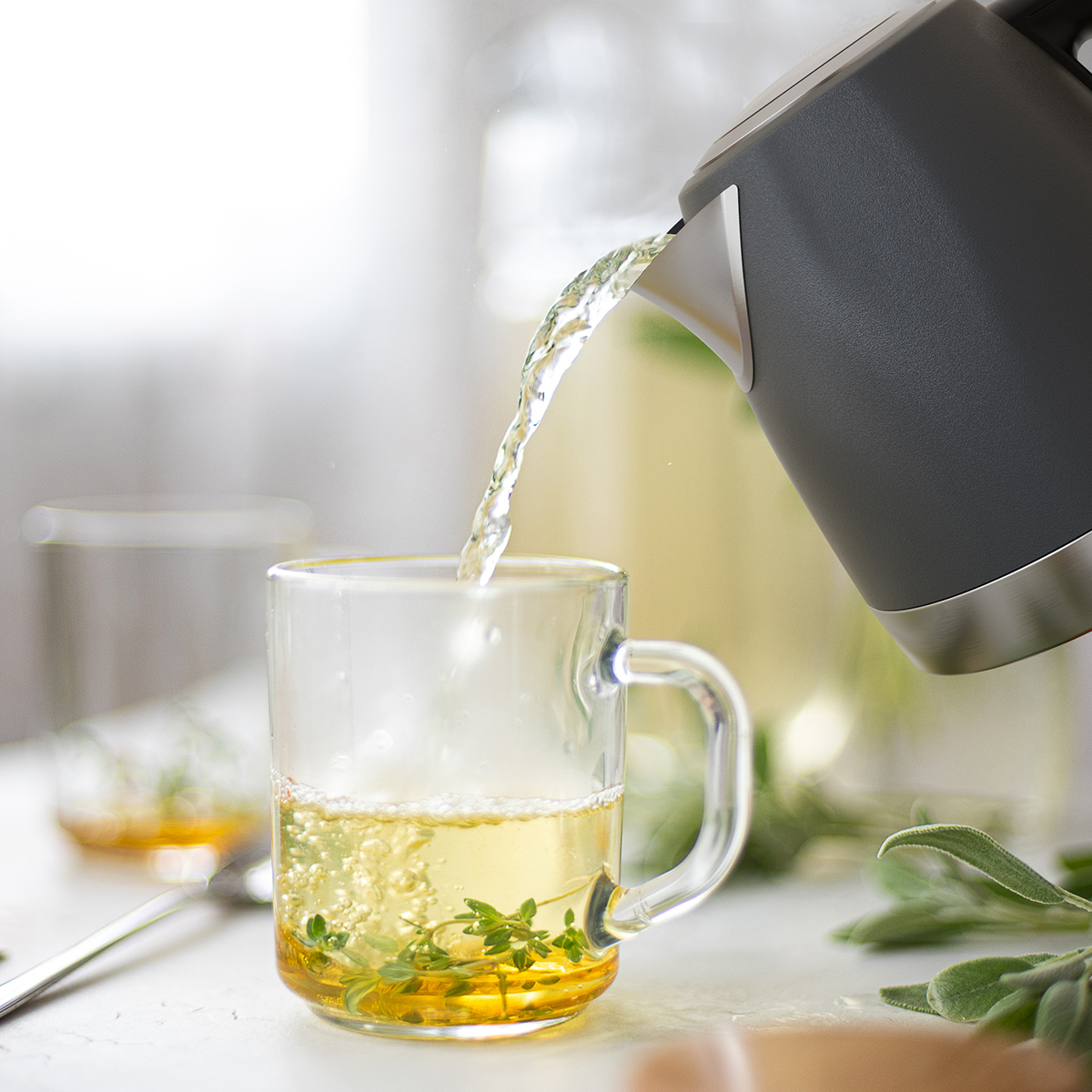 De l'eau chaude est versée d'une bouilloire grise dans une tasse en verre avec des herbes.