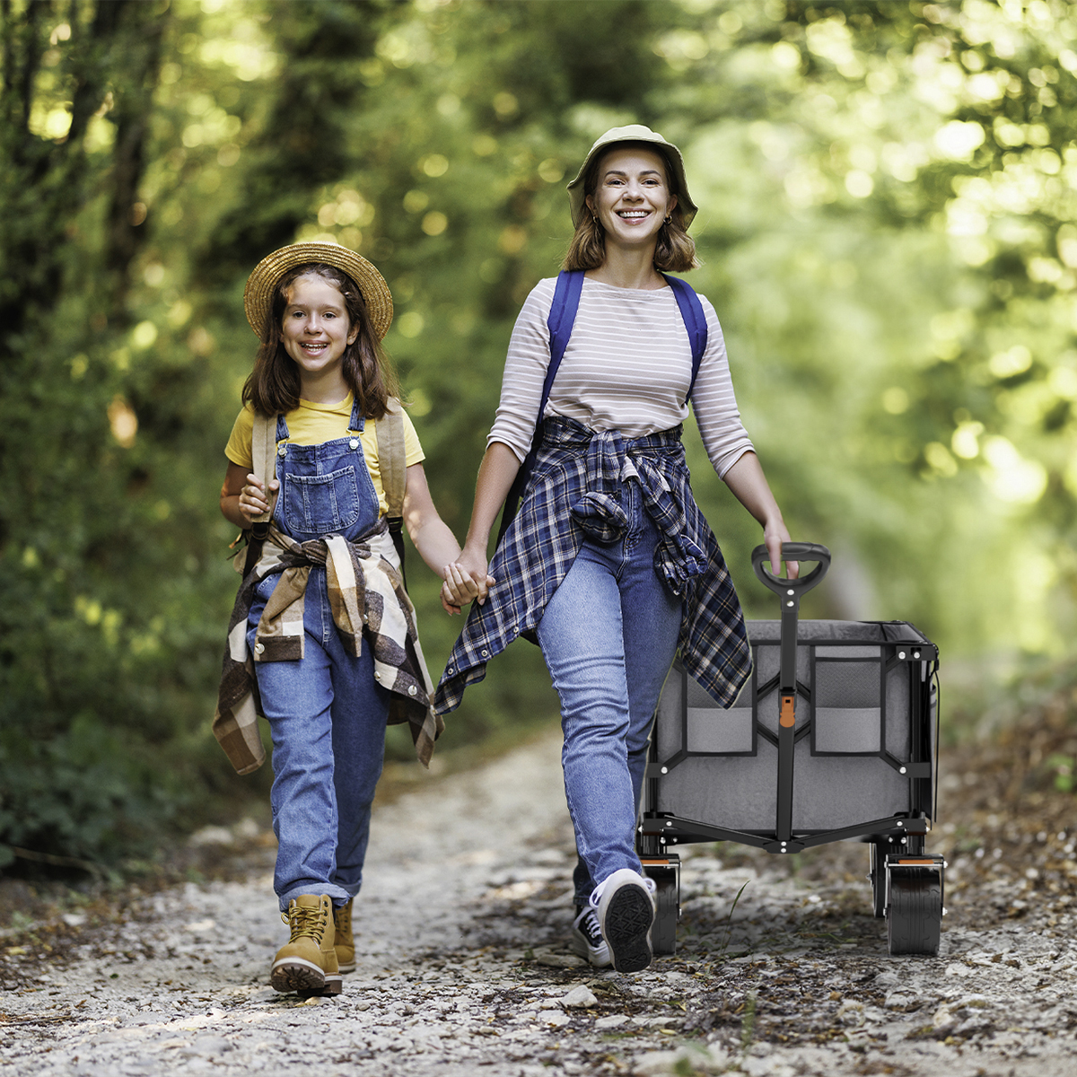 Two women walk on a forest path, one pulling a gray wagon.