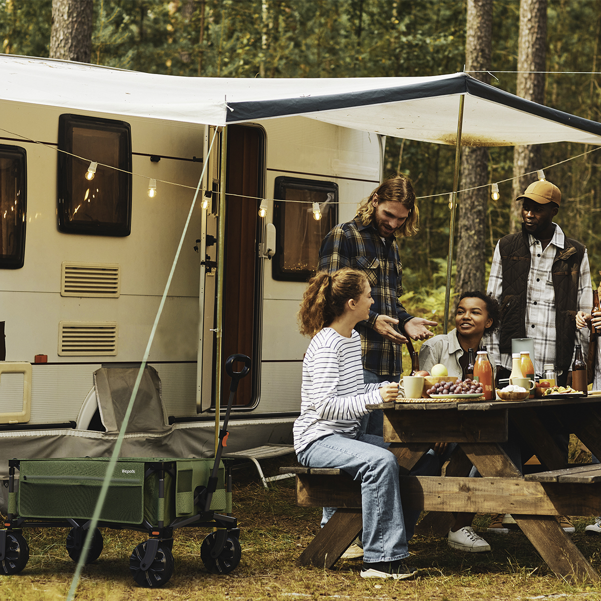 People are camping next to a caravan, with a picnic table full of food and drinks.