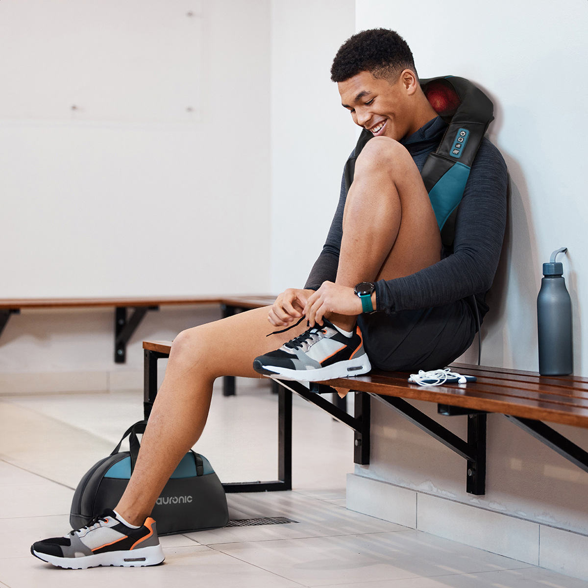 A man sits on a locker room bench tying his shoes. He wears a neck massager.