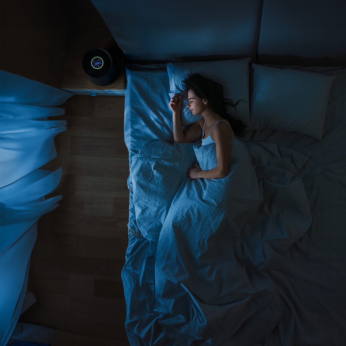 Woman sleeping in bed, air purifier on nightstand, blue lighting.