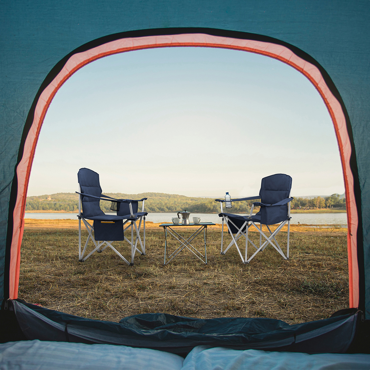 Two blue camping chairs and a table with a coffee pot by a lake, viewed from inside a tent.