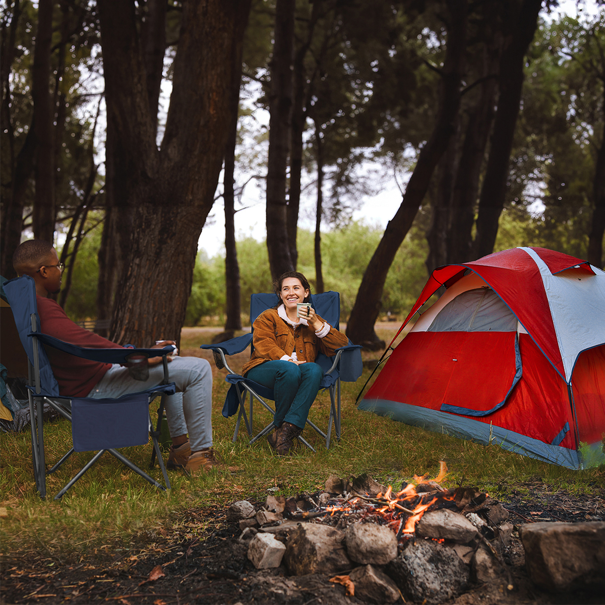 Two people sit in camping chairs next to a red tent by a campfire in the woods.