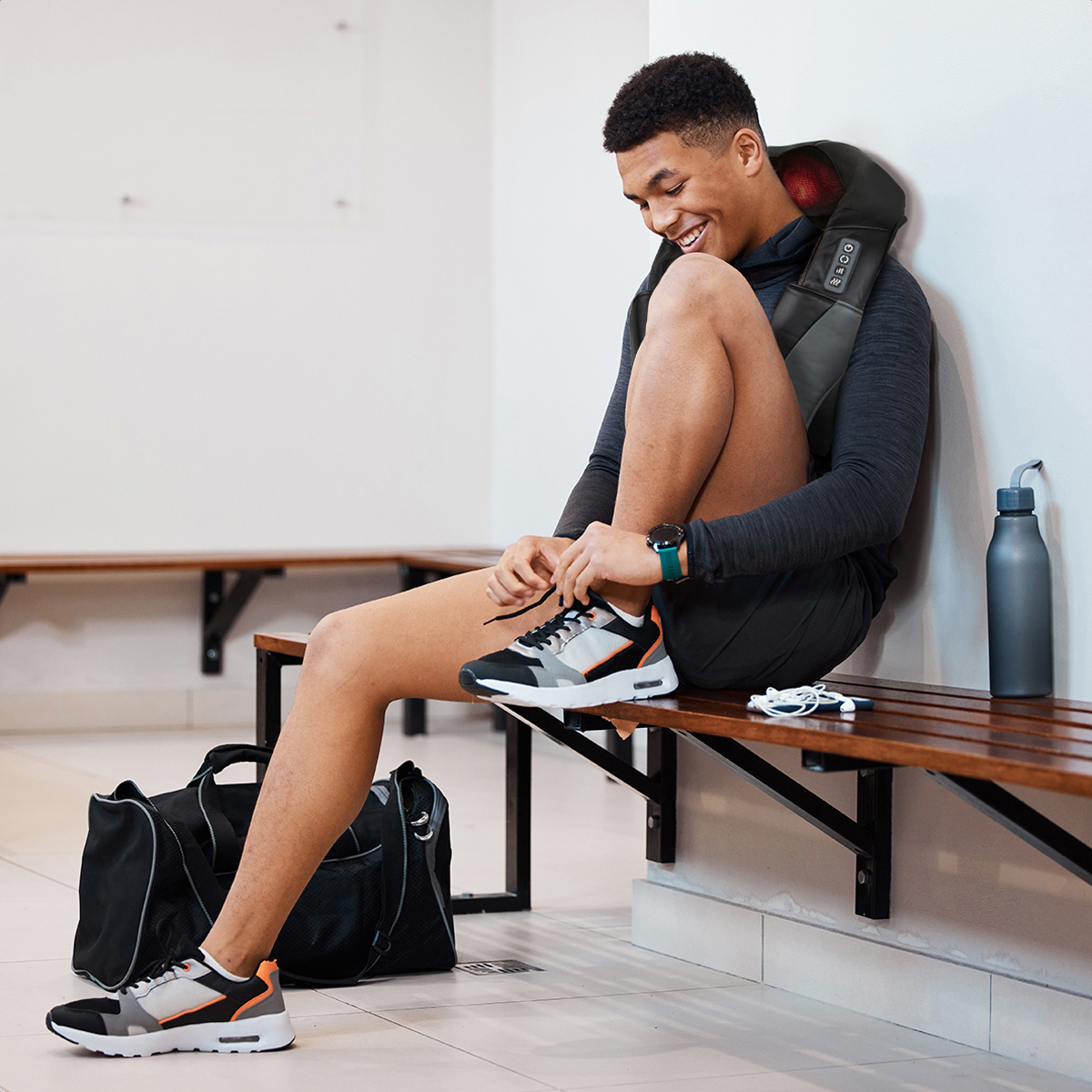 A man with a neck massager ties his sneakers on a wooden bench.