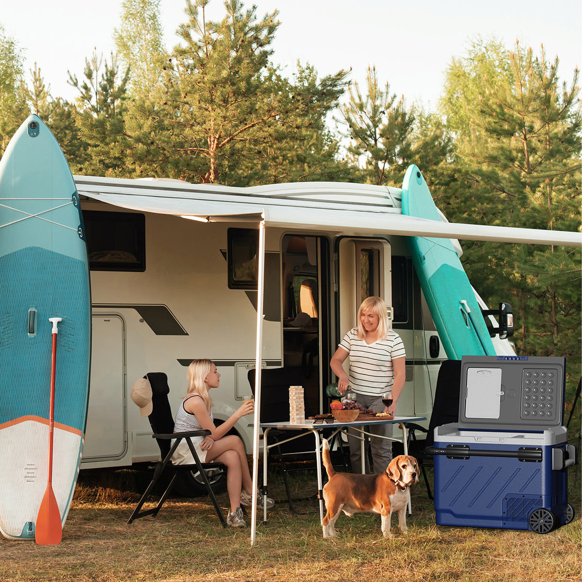 Twee vrouwen en een hond kamperen met een camper; twee surfplanken en een koelbox zijn aanwezig.