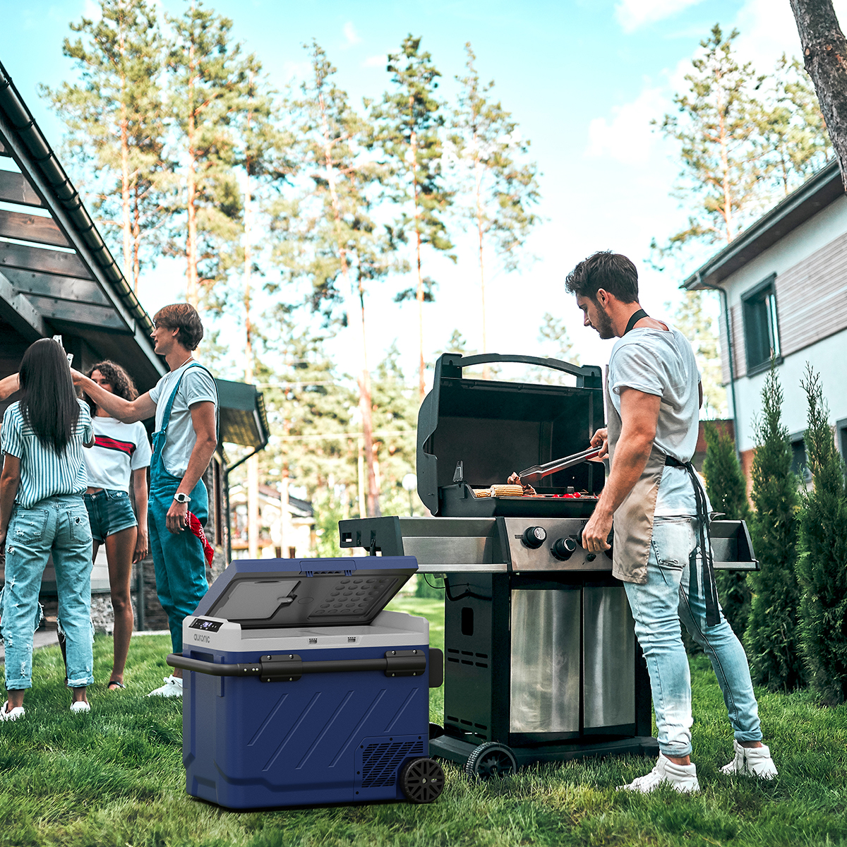 Mensen verzamelen zich voor een barbecue in de buitenlucht. Een man grilt eten, terwijl anderen bij een blauwe koelbox kletsen.