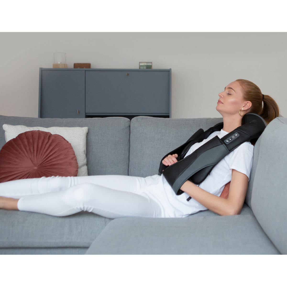 Woman on gray sofa with neck massager, eyes closed.