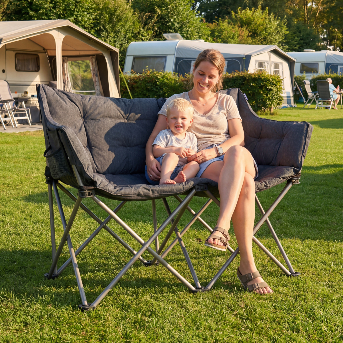Woman with child sitting on a dark camping chair on grass in front of caravans.