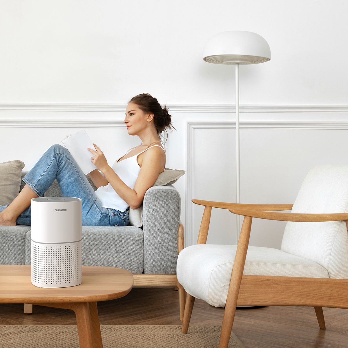 Woman reading on sofa next to air purifier. White armchair and floor lamp.