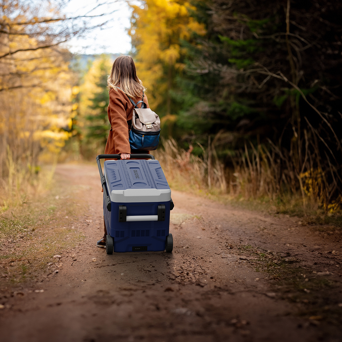 Een vrouw met rugzak trekt een blauwe koelbox over een landweg in de herfst.