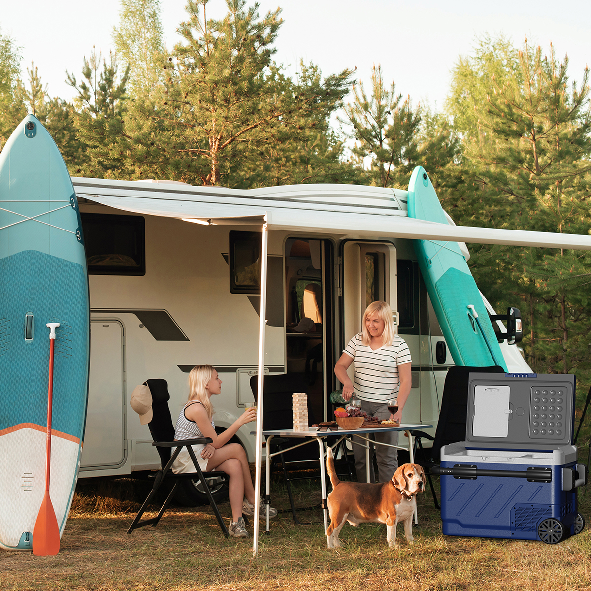 Twee vrouwen en een hond kamperen bij een camper, omringd door bomen.