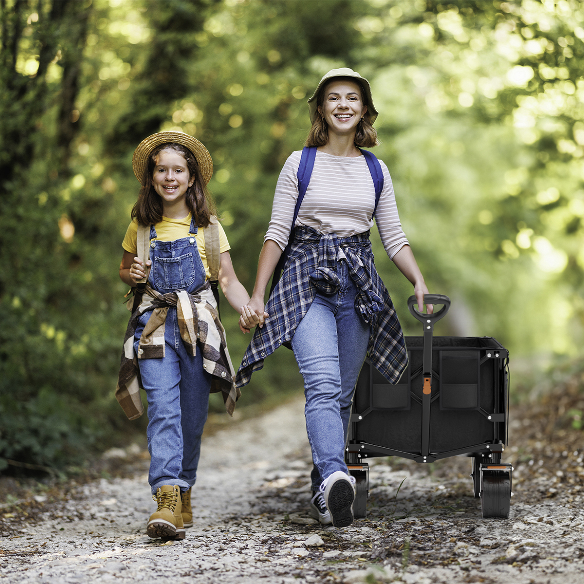 Two women walk hand in hand on a forest path, pulling a black wagon.