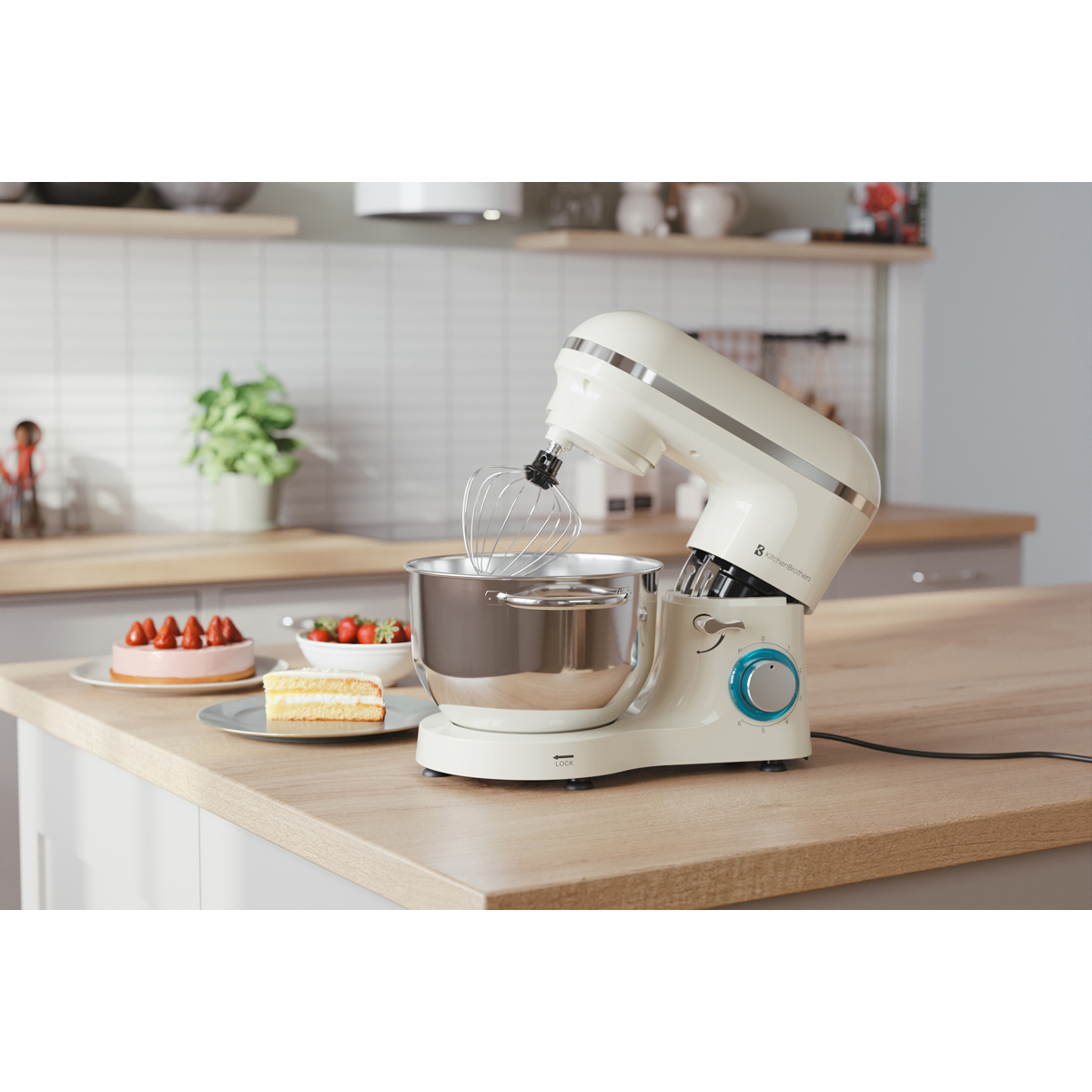 Een crème-kleurige standmixer met garde staat op een houten aanrecht met taart en aardbeien.