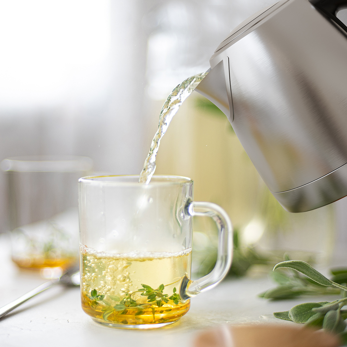 Heißes Wasser wird in eine Tasse mit Kräutern gegossen. Ein Wasserkocher ist zu sehen.