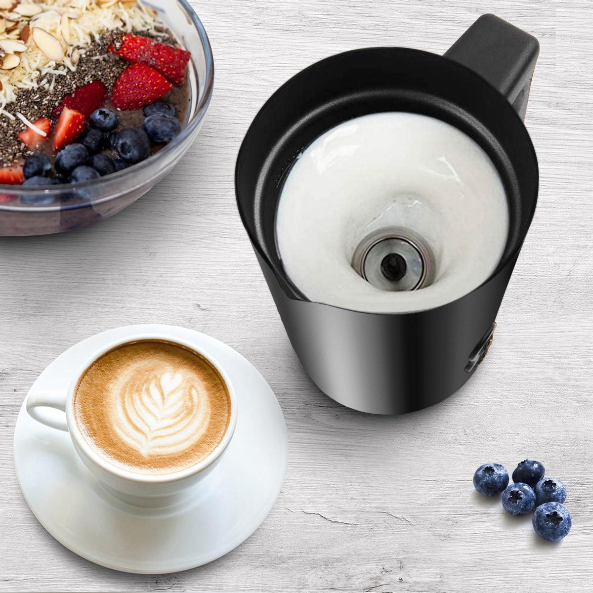 Schwarzer Milchaufschäumer mit weißem Schaum, daneben eine Kaffeetasse und Beeren.