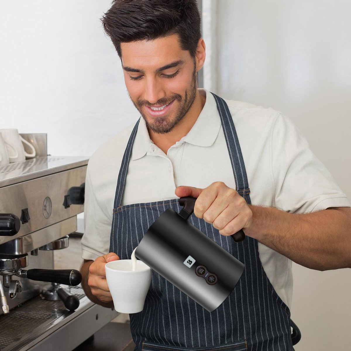 Ein Mann gießt Milch aus einem schwarzen Milchaufschäumer in eine weiße Tasse neben einer Kaffeemaschine.