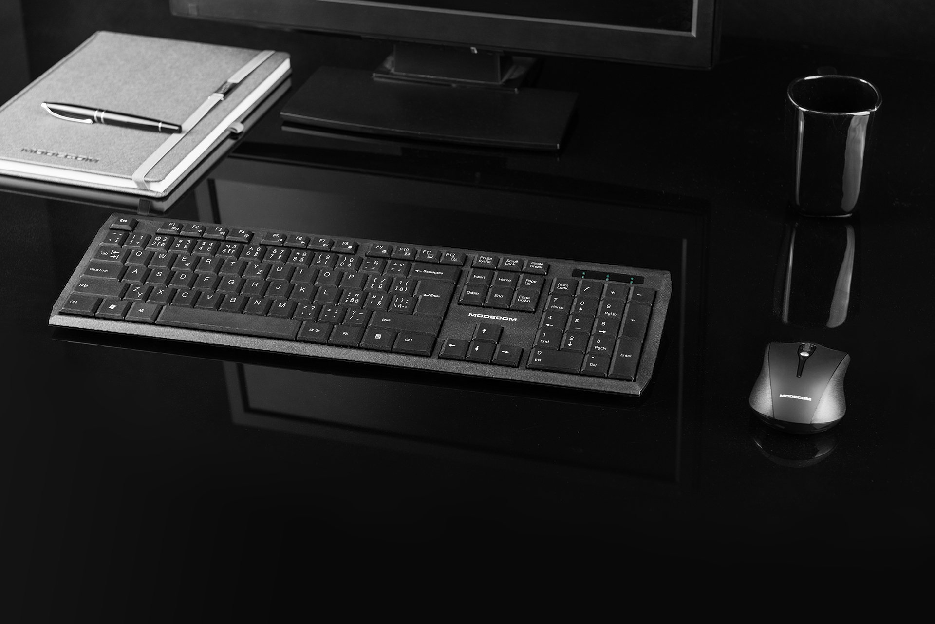 Czarne biurko z klawiaturą, myszką, notatnikiem, długopisem i kubkiem. Monitor komputera w tle.