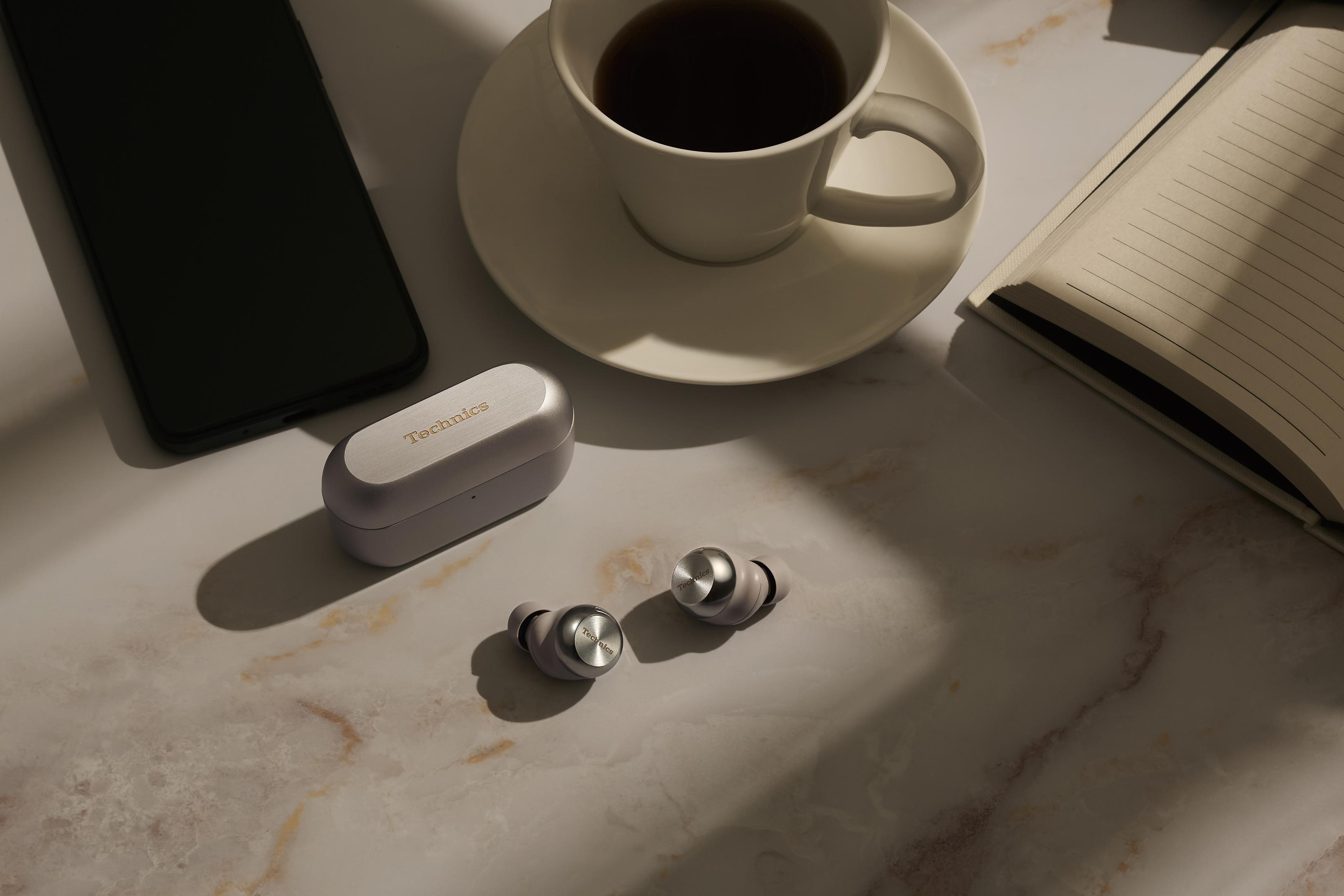 Ein Smartphone, Kopfhörer mit Ladebox, Kaffeetasse und Notizbuch auf Marmor.