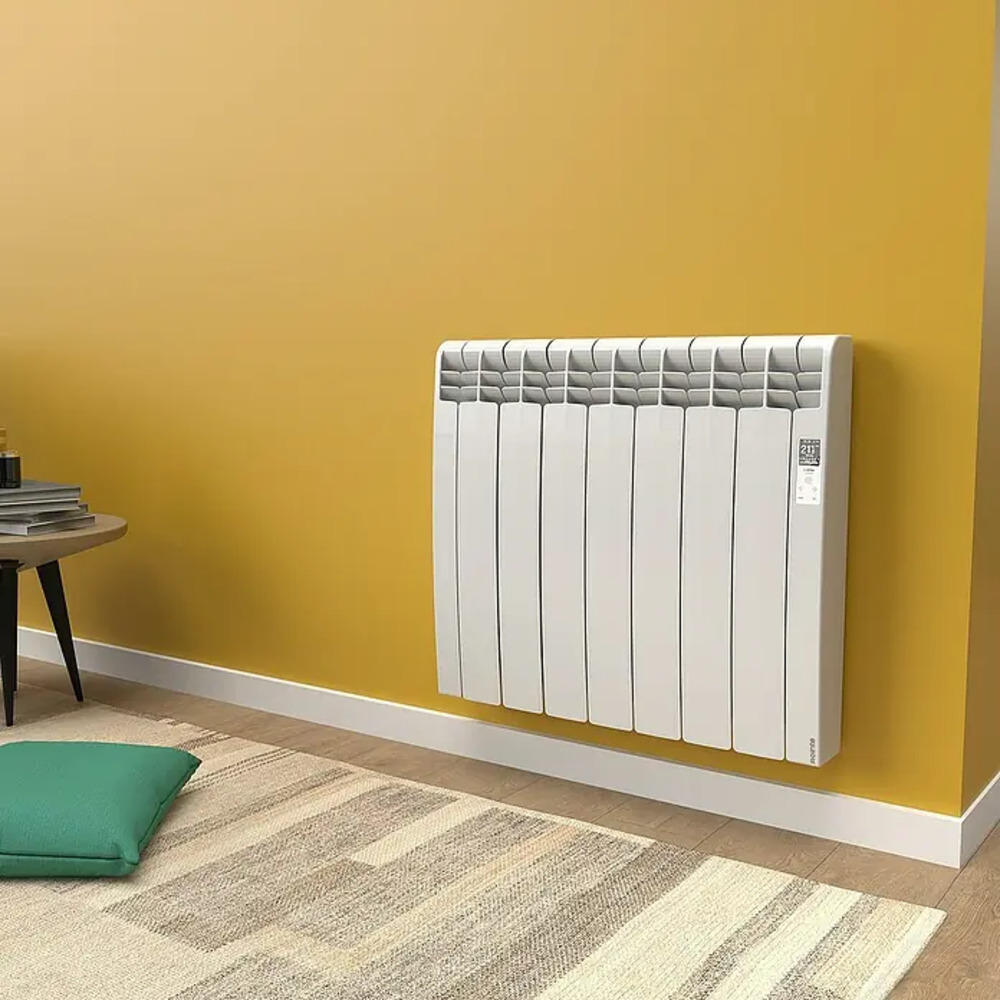 Un radiador blanco montado en una pared amarilla, con alfombra y cojín delante.