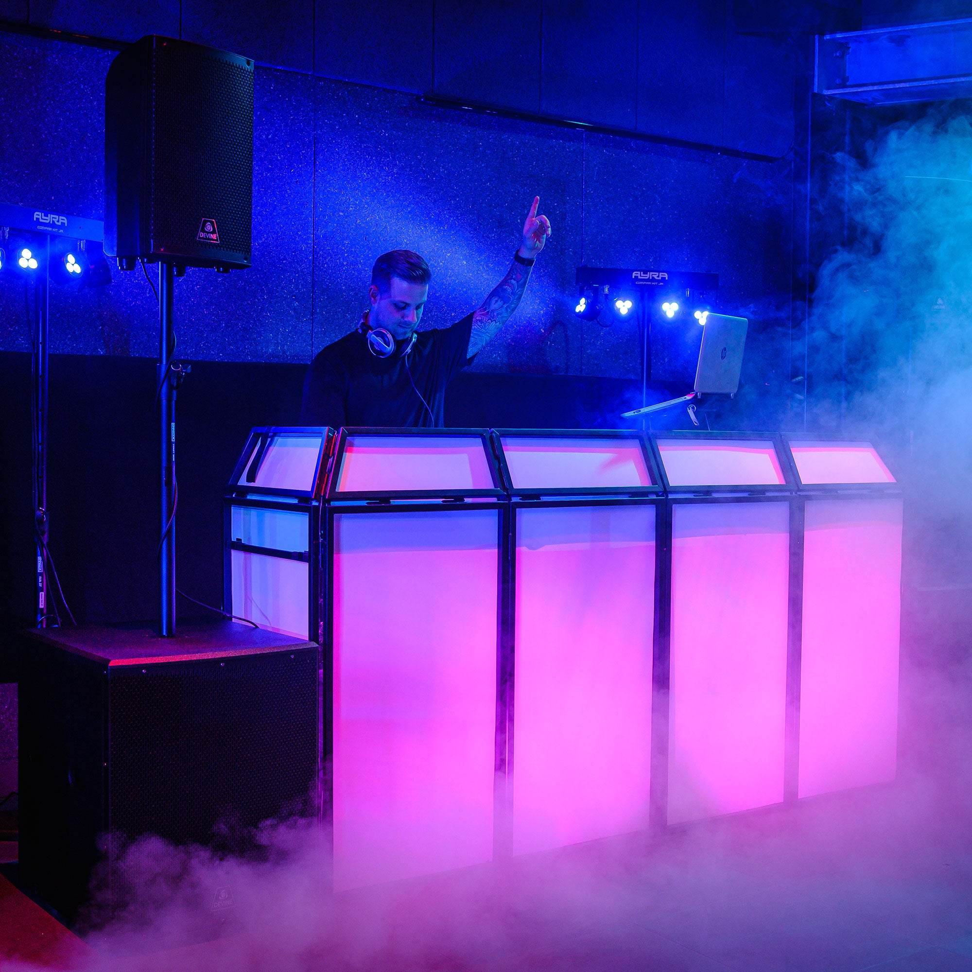Een DJ met koptelefoon staat achter een verlichte console, één hand omhoog.