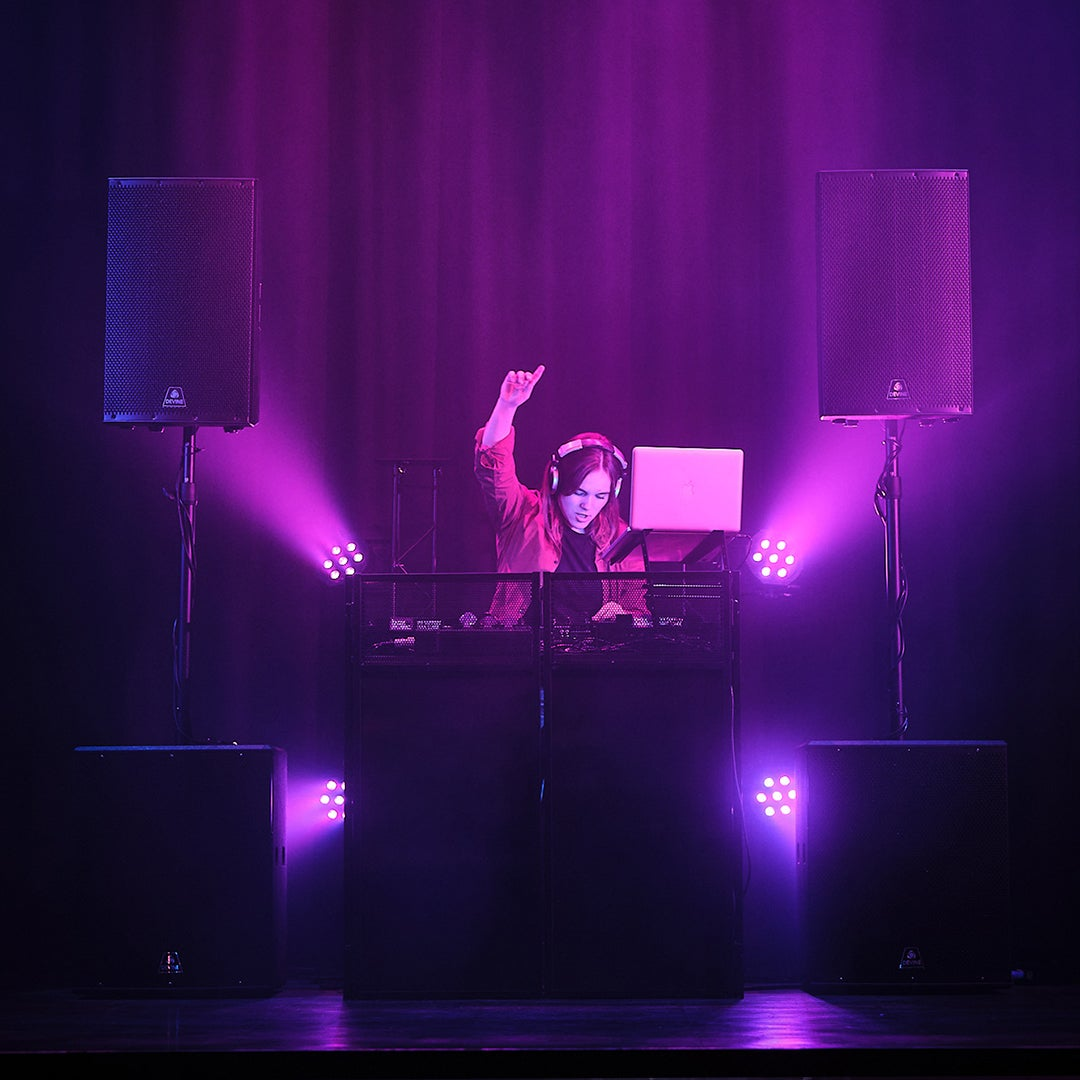 DJ met koptelefoon op het podium, verlicht door paarse lampen, met luidsprekers.