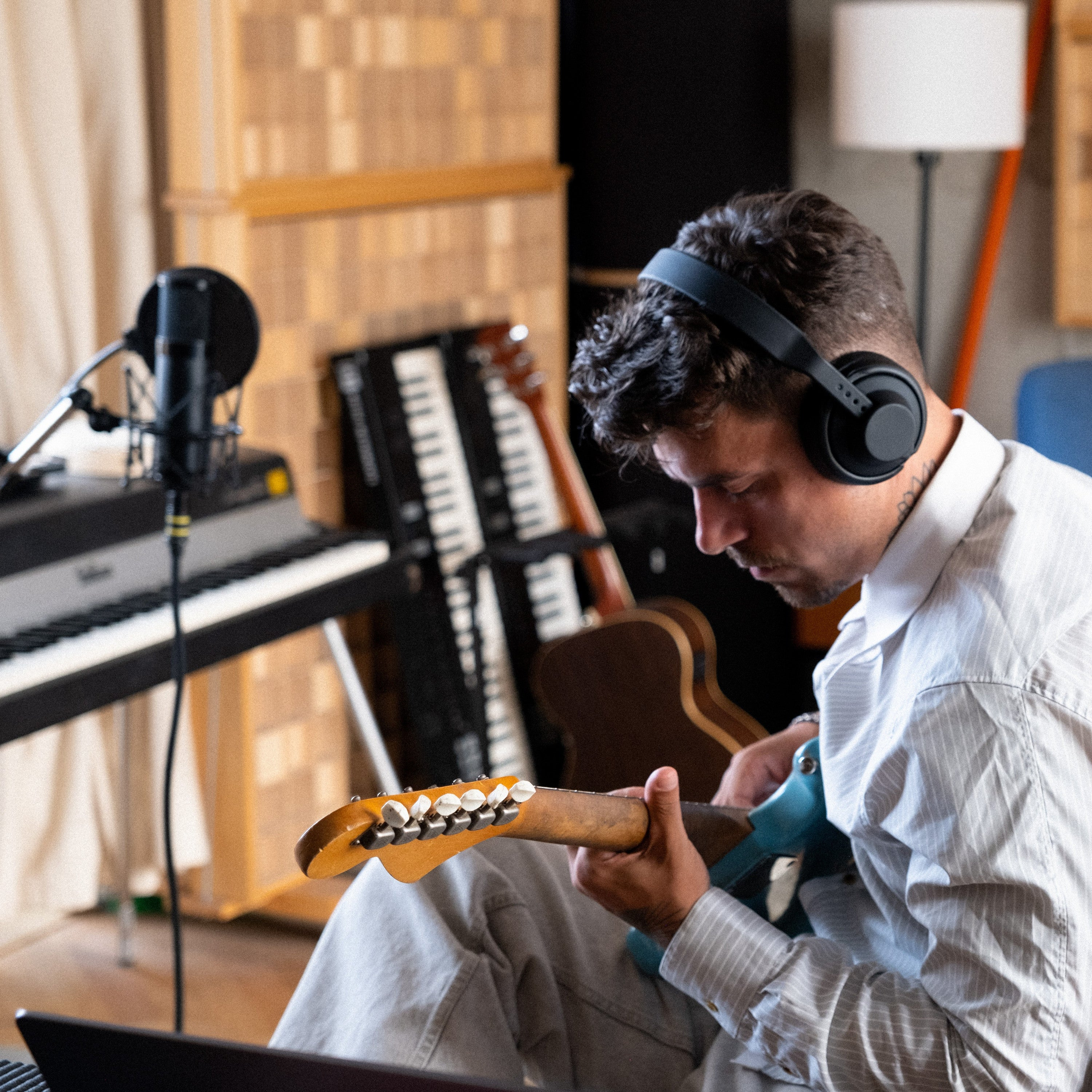 Man met koptelefoon speelt lichtblauwe gitaar in studio met keyboards en microfoon.