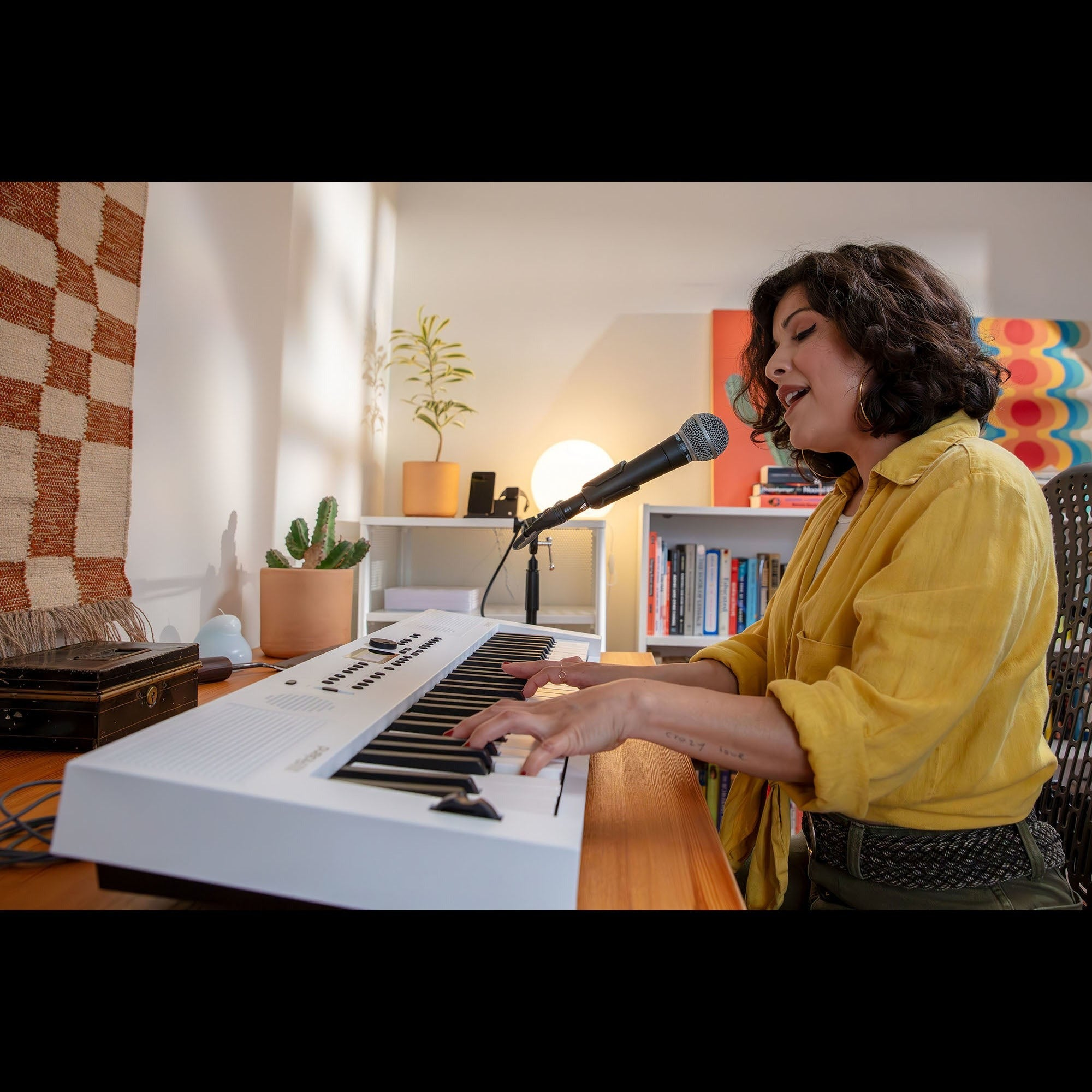 Een vrouw speelt keyboard en zingt in een microfoon, met boeken en planten op de achtergrond.
