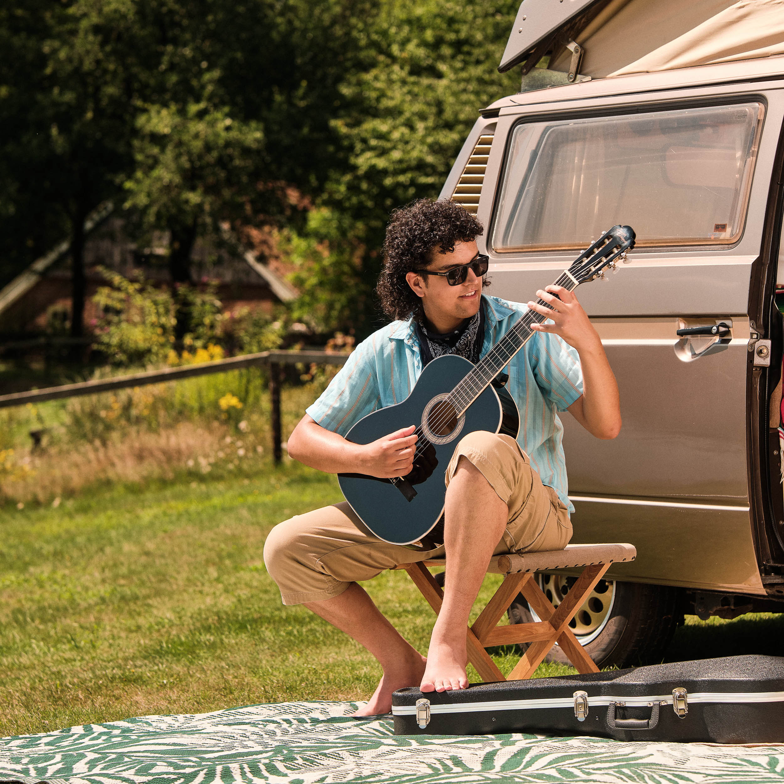Een man met krullend haar speelt gitaar naast een camper op een deken.