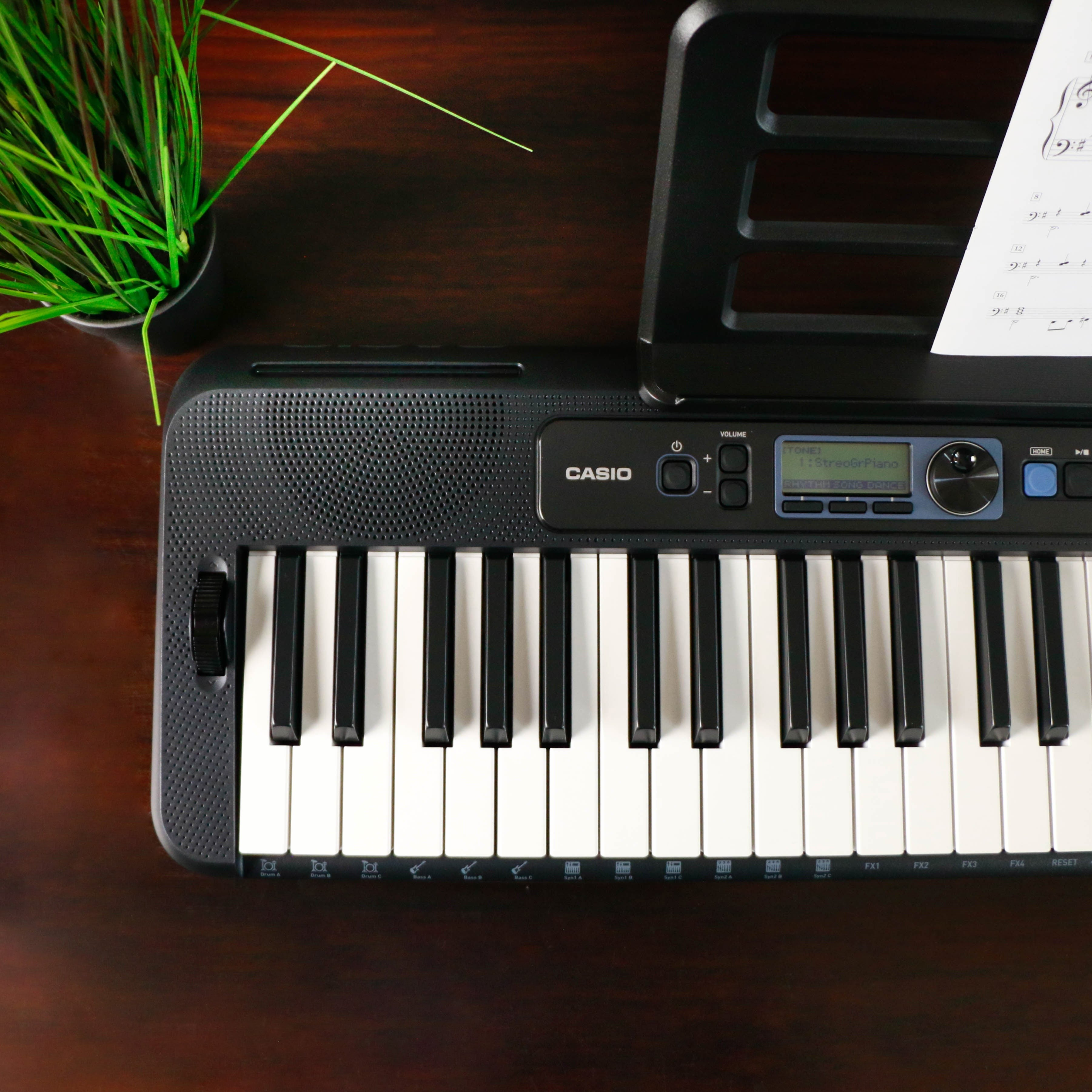 Een zwart Casio keyboard met bladmuziek en plant op bruine houten tafel.