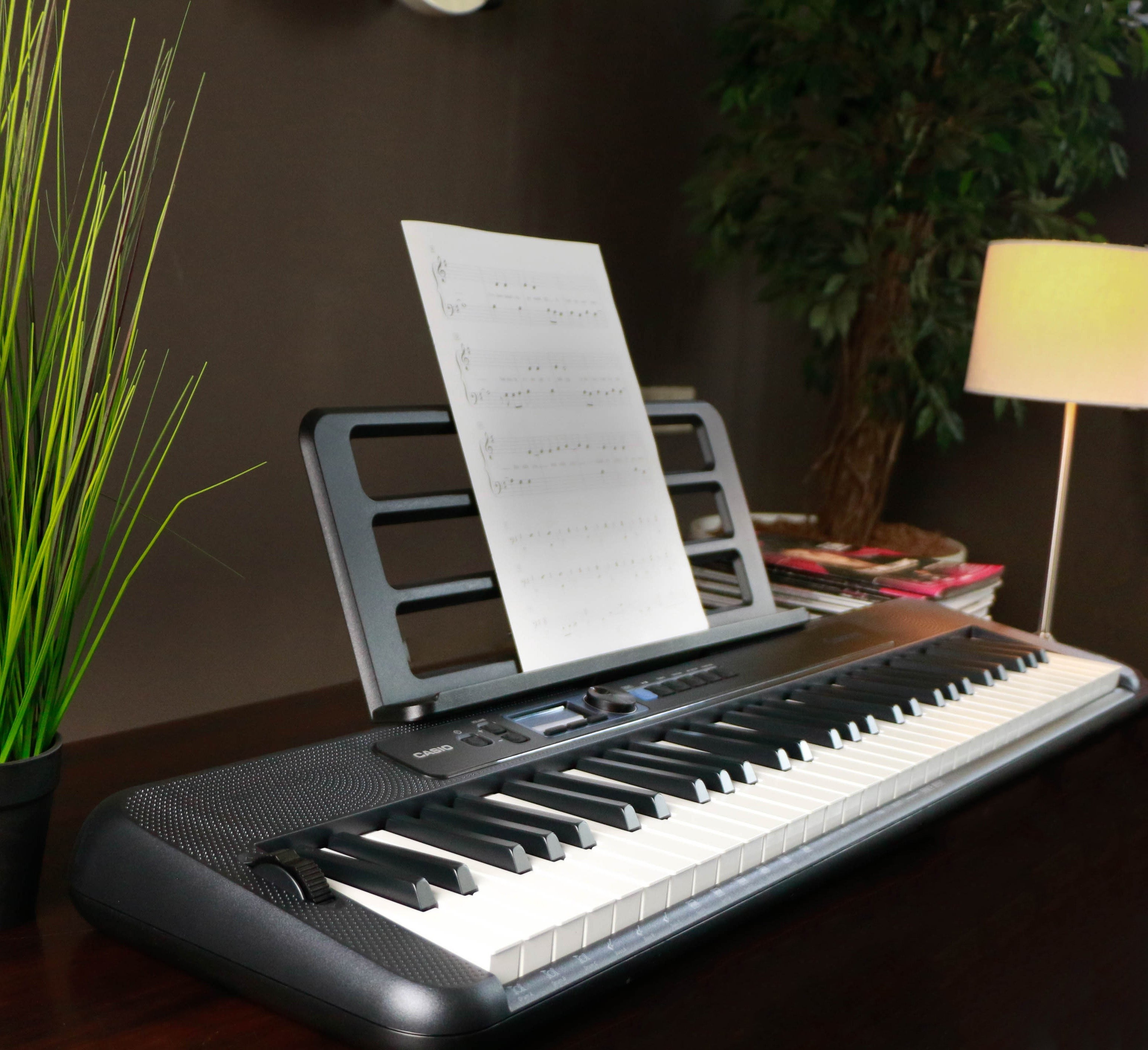 Een zwart Casio-keyboard met bladmuziek op een standaard, naast een plant en een lamp.