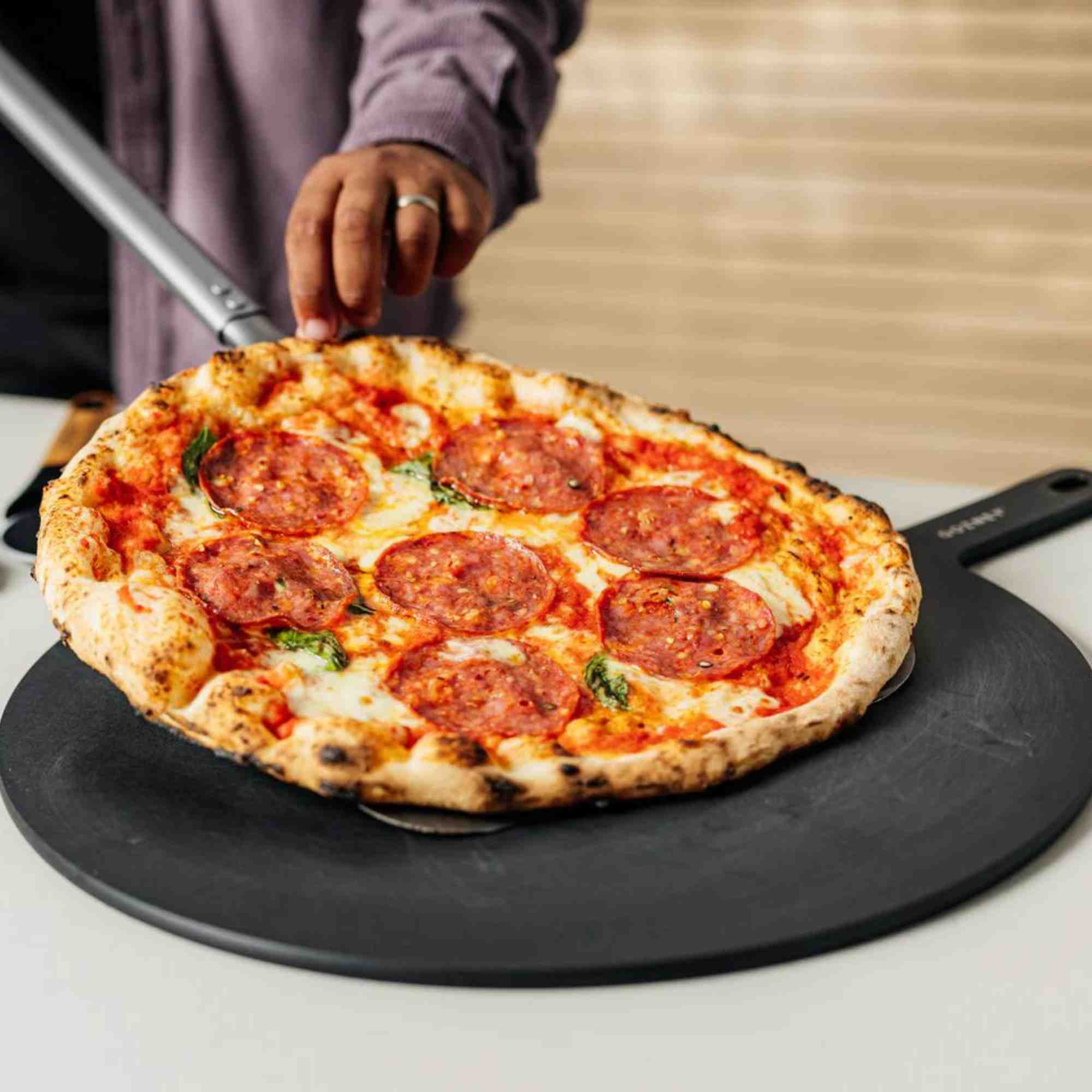 Eine Person schiebt eine Salamipizza mit einem Pizzaschieber von einer schwarzen Platte.