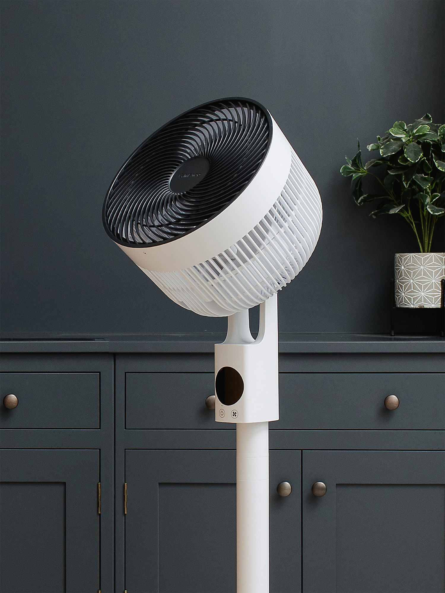 Ein weißer Standventilator mit schwarzem Gitter steht vor einem dunkelgrauen Schrank.
