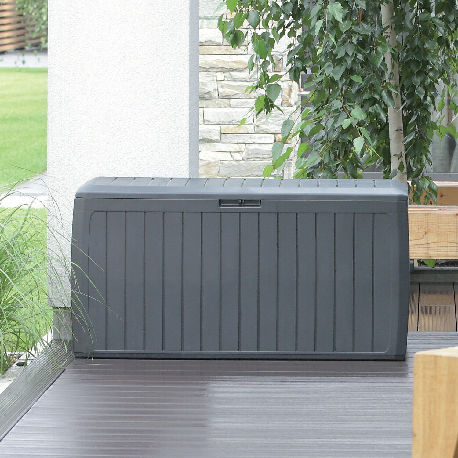 Una caja de almacenamiento gris oscuro sobre una terraza con un abedul y muro de piedra detrás.