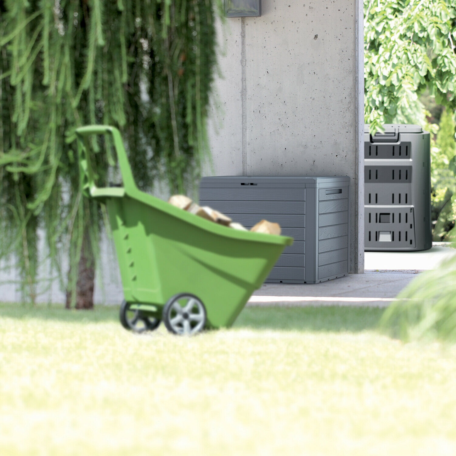 Carretilla verde cargada de leña, al lado una caja de jardín gris y un compostador.