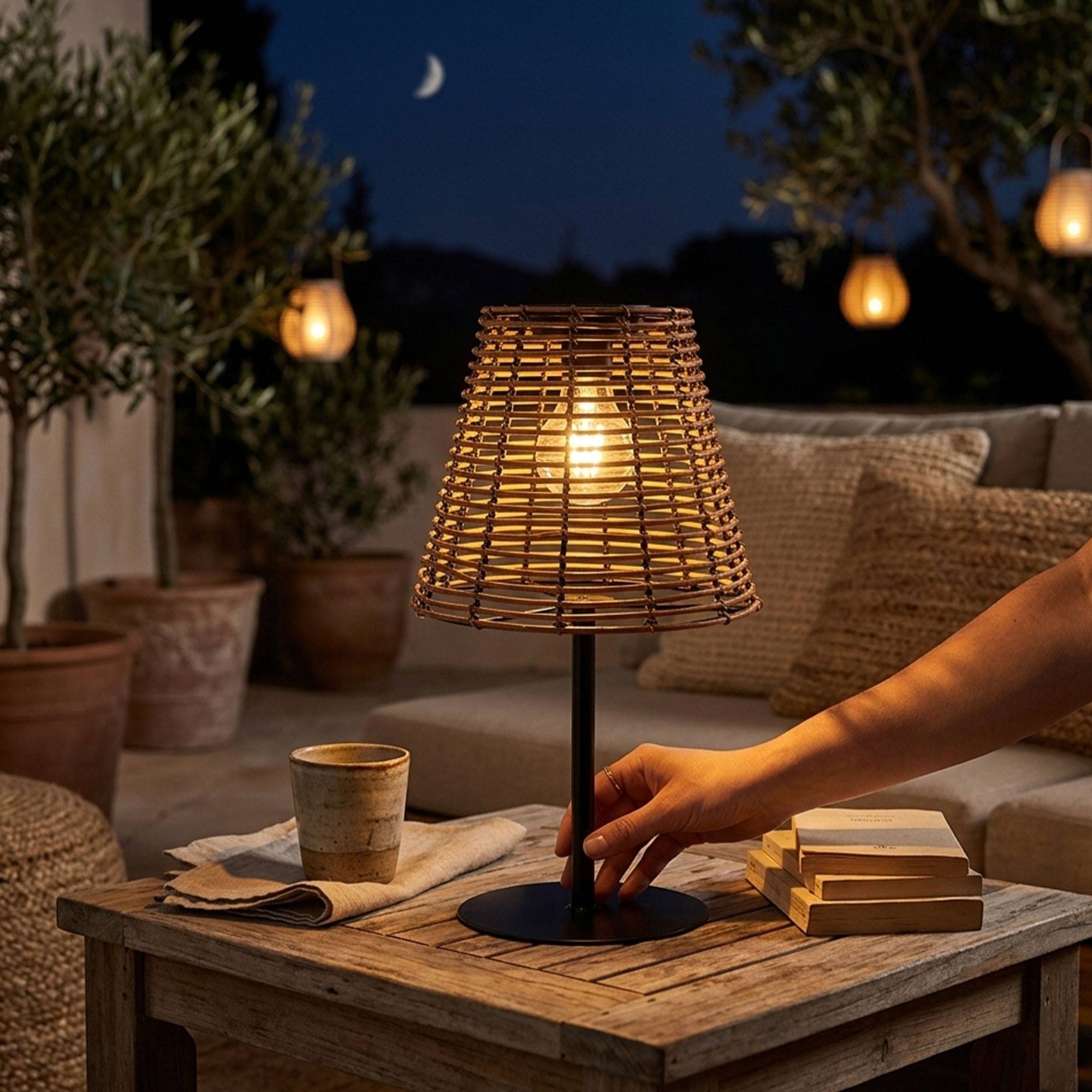 Lámpara de mimbre en mesa de madera de noche, mano alcanzándola. Exterior.