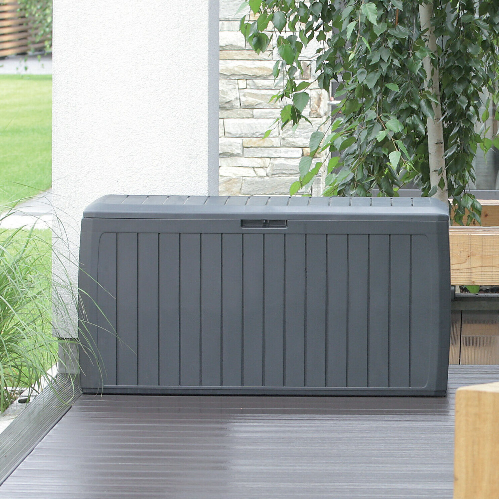 Una caja de almacenamiento gris se asienta sobre una terraza de madera cerca de hierba verde y árboles.