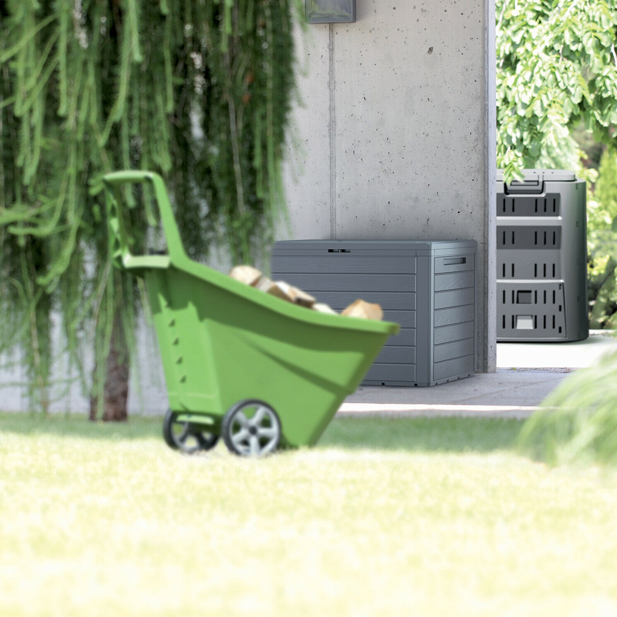 Un carrito de jardín verde cargado de leña, junto a cajas de almacenamiento grises.