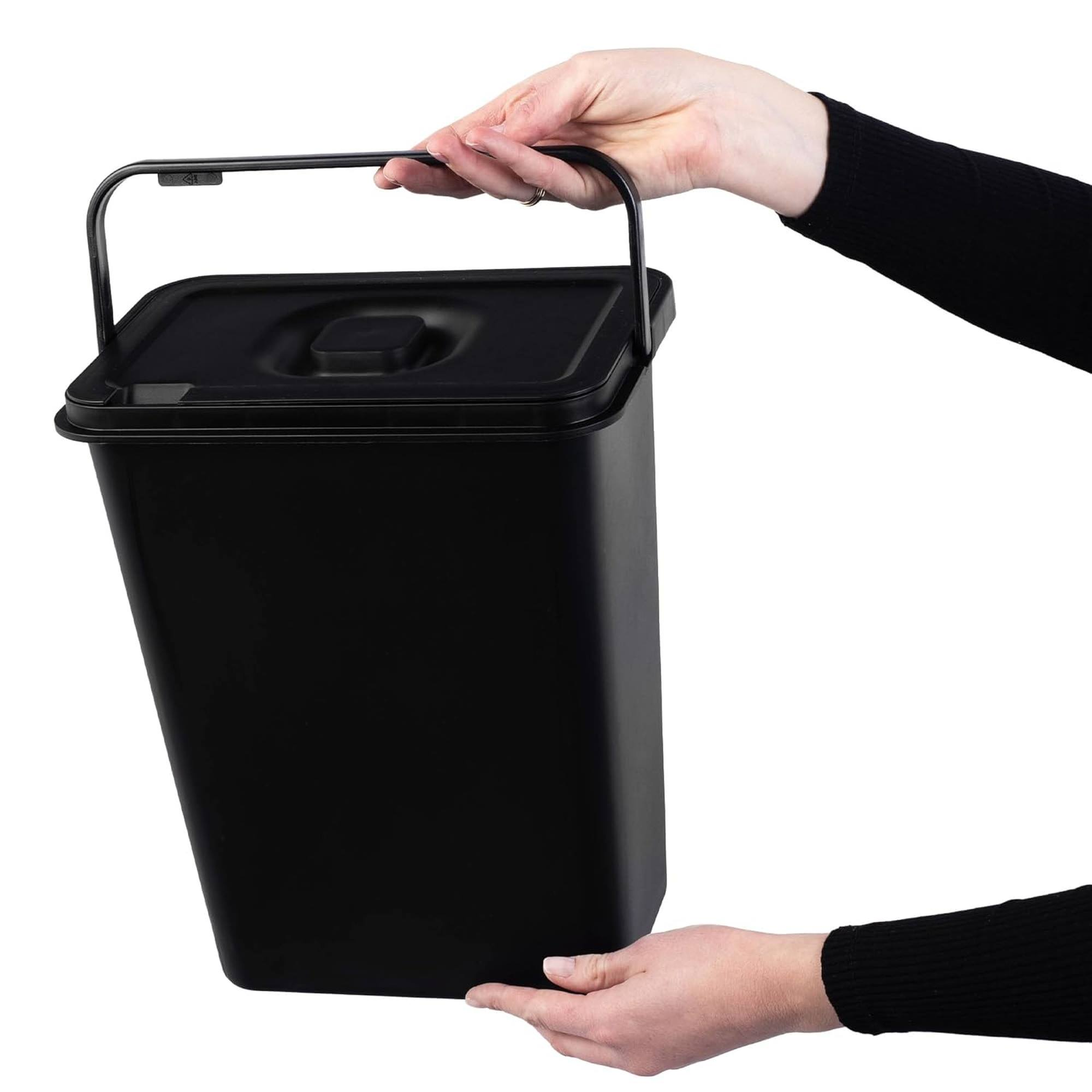 Cubo de basura negro con asa y tapa, sostenido por dos manos.