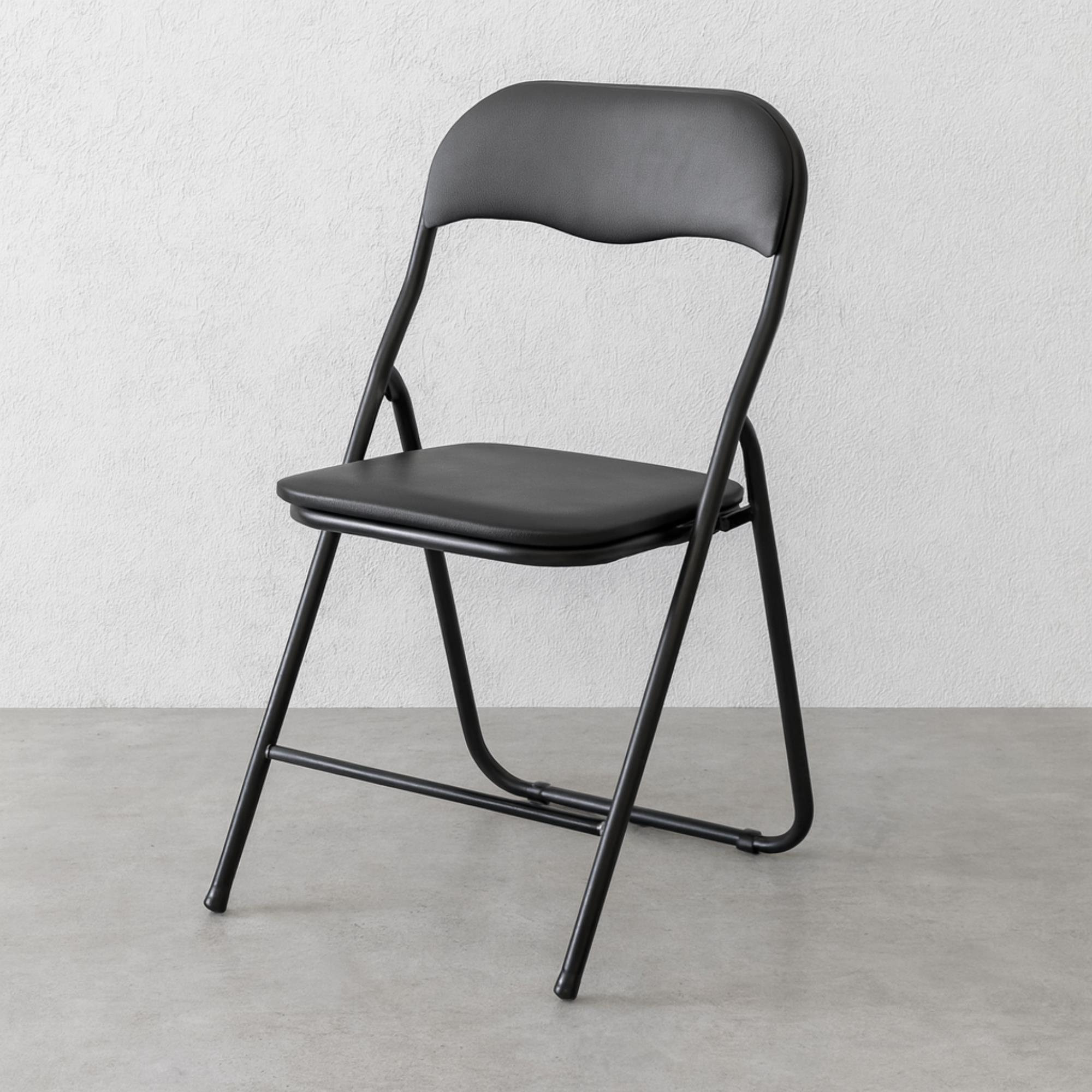 Una silla plegable negra sobre suelo de hormigón gris contra pared blanca.
