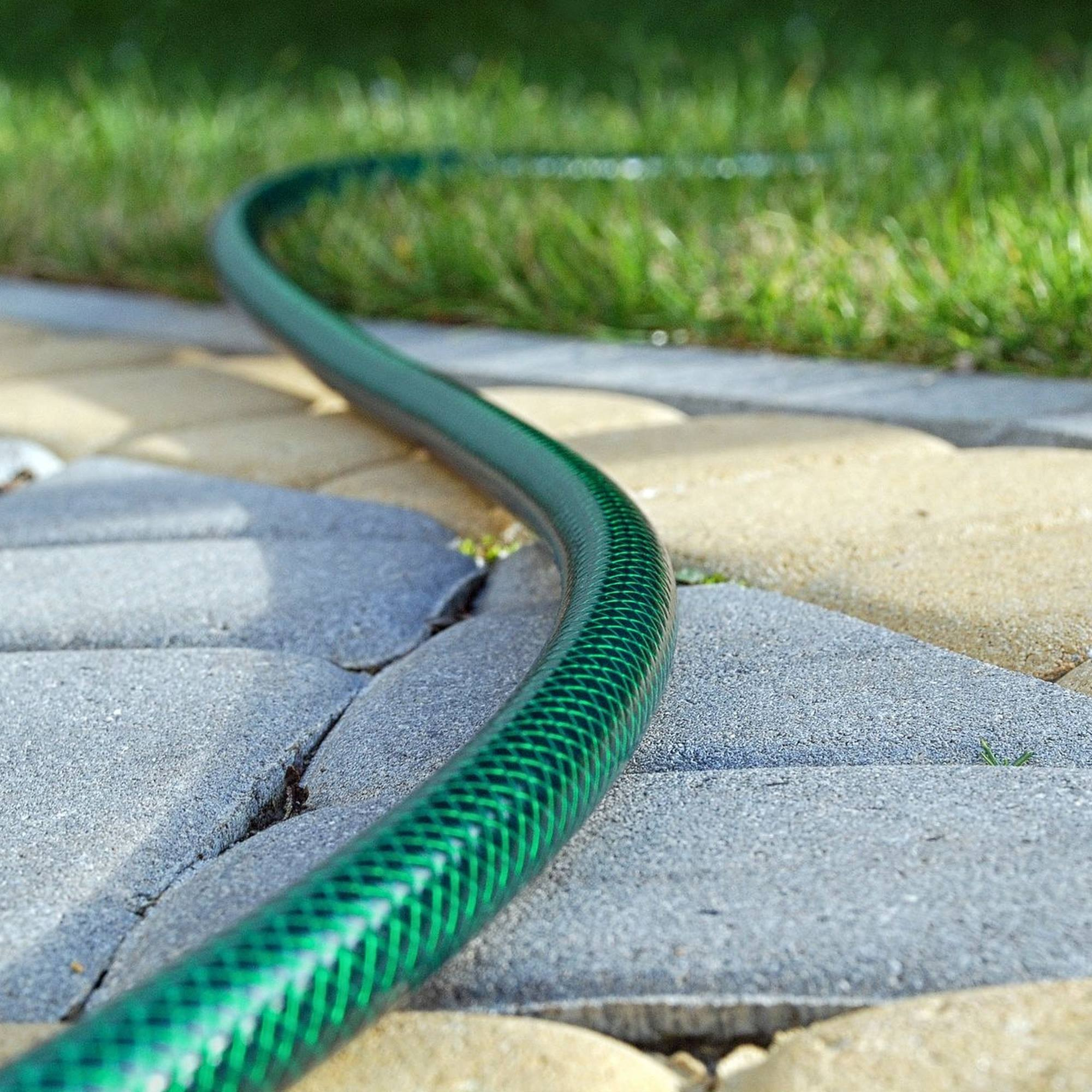Ein grüner Gartenschlauch liegt auf grauen und beigen Pflastersteinen neben grünem Gras.