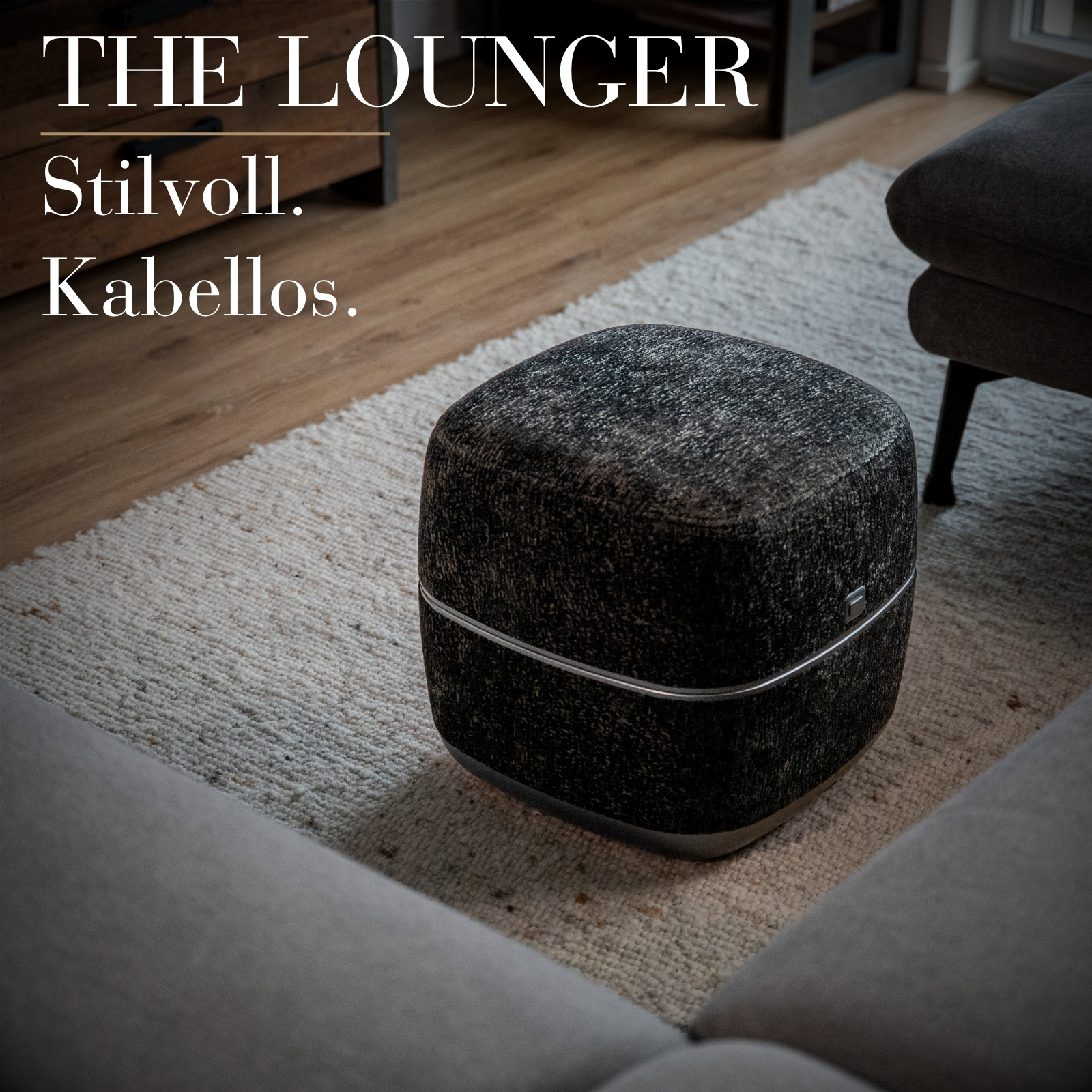 Ein dunkler, quaderförmiger Lautsprecher steht auf einem hellen Teppich neben einer Couch.