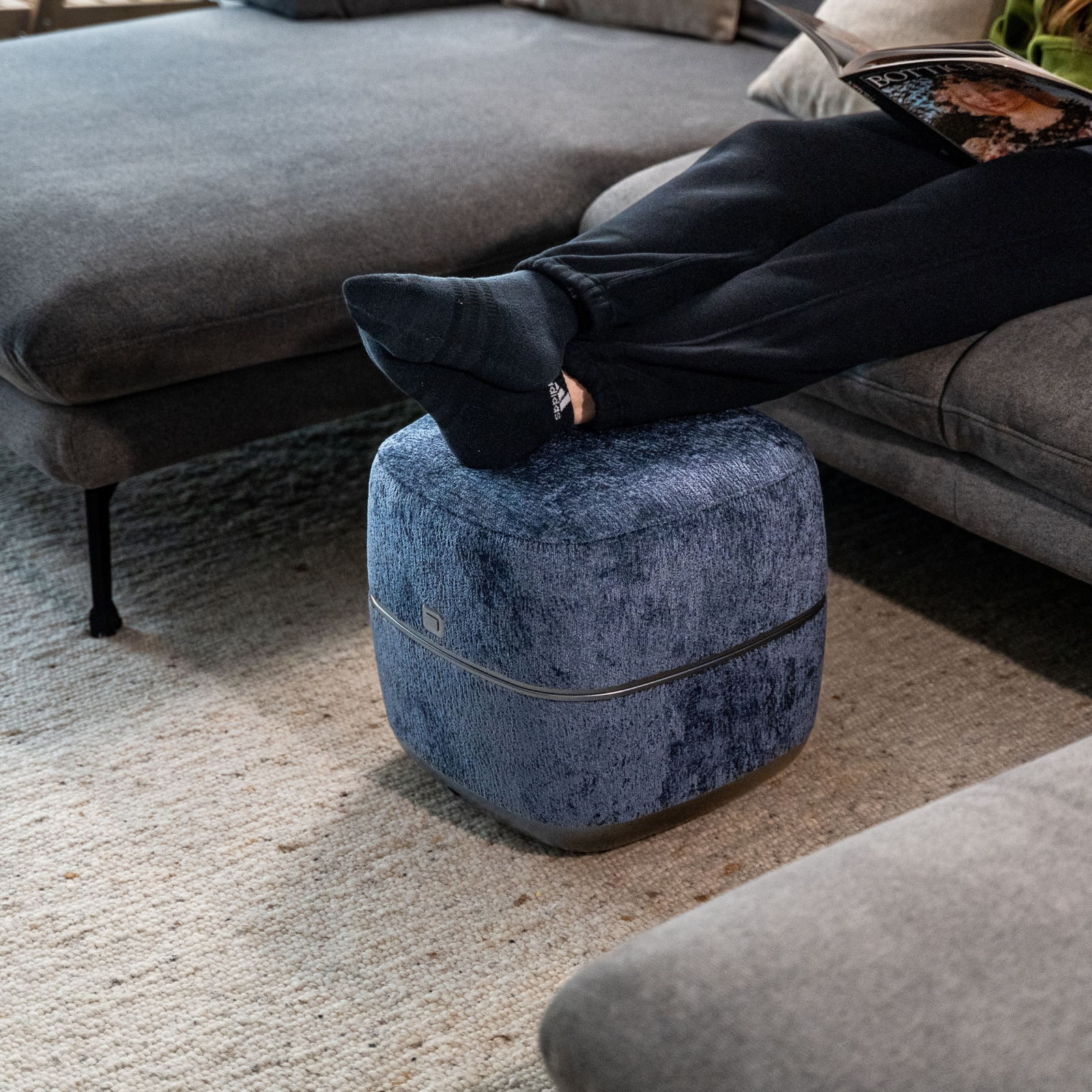Ein blauer Hocker steht auf einem Teppich vor einem Sofa. Ein Fuß ruht darauf.