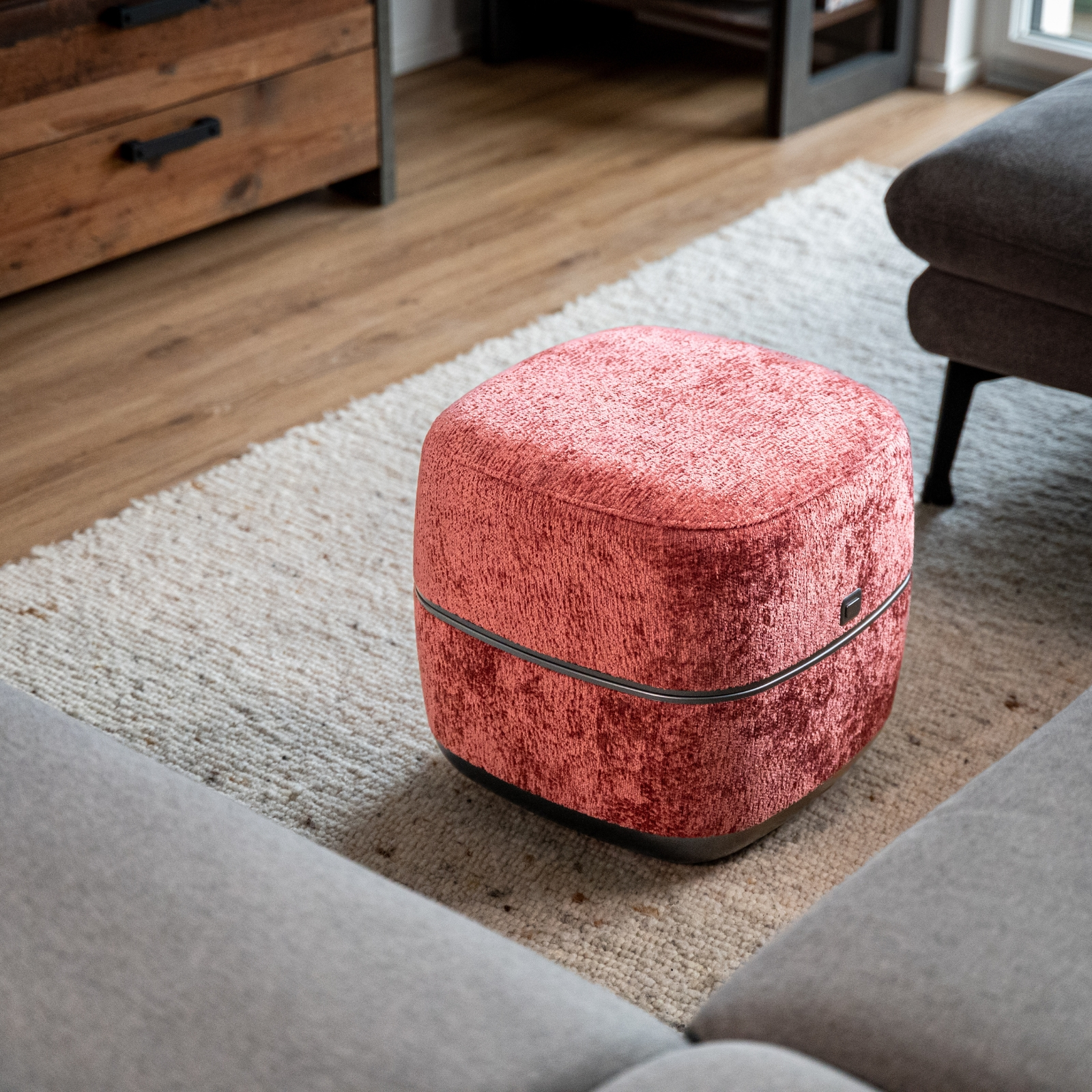 Ein korallenroter Hocker mit Samtbezug steht auf einem Teppich neben einem grauen Sofa.