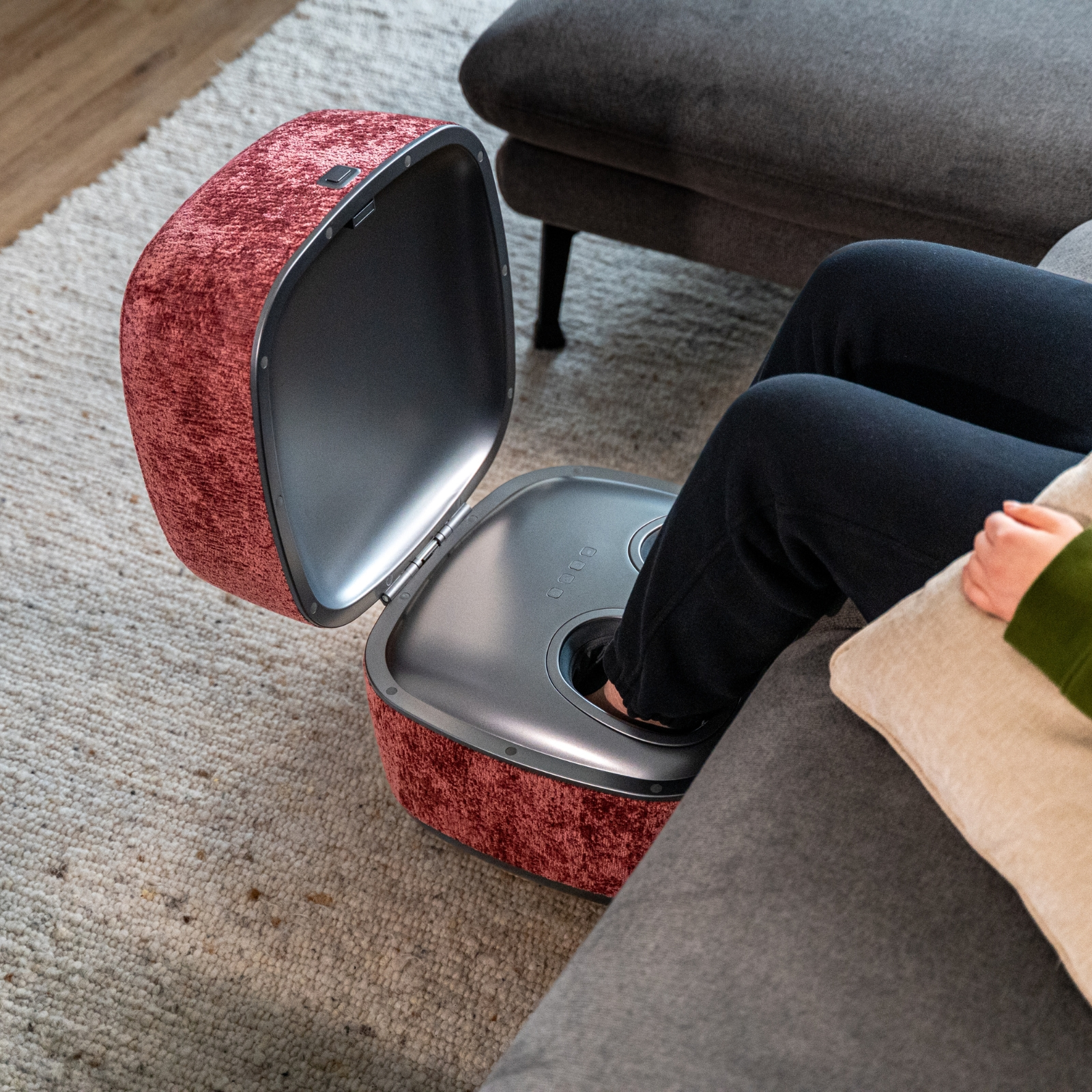 Eine Person ruht ihre Füße in einem roten Samt-Fußmassagegerät neben einem grauen Sofa.